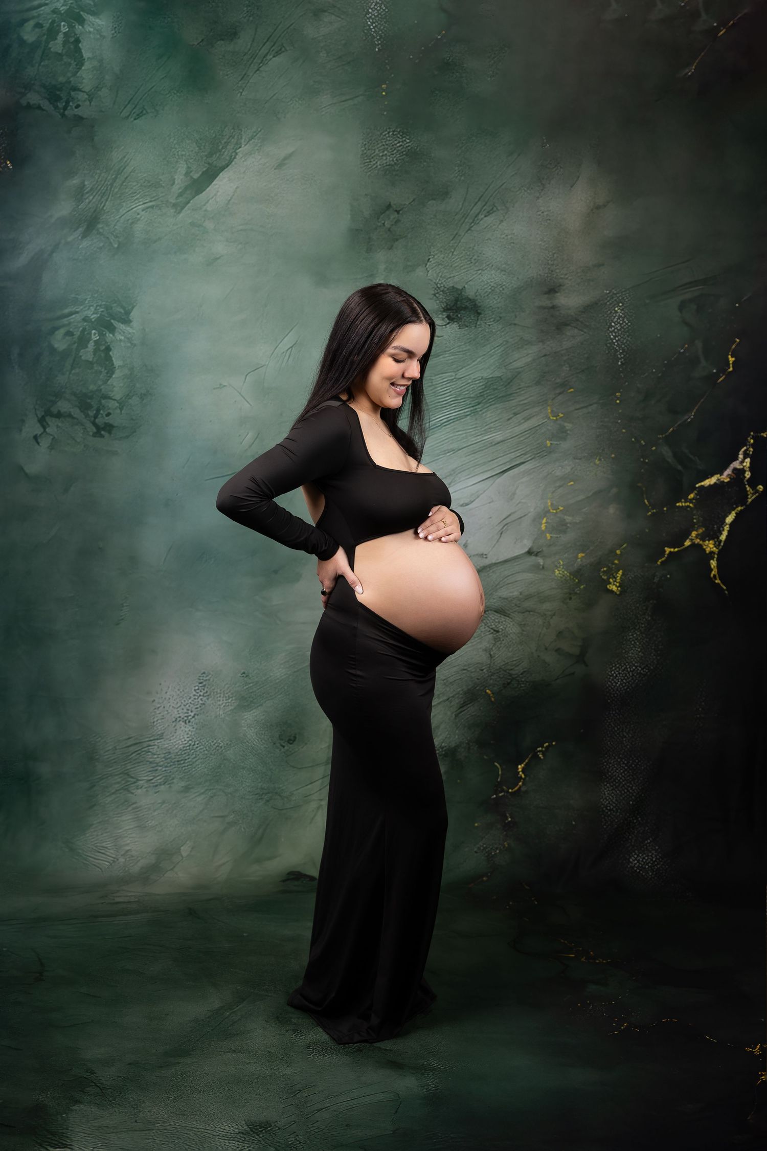 Pregnant woman in black dress cradling baby bump against a dark green textured backdrop.