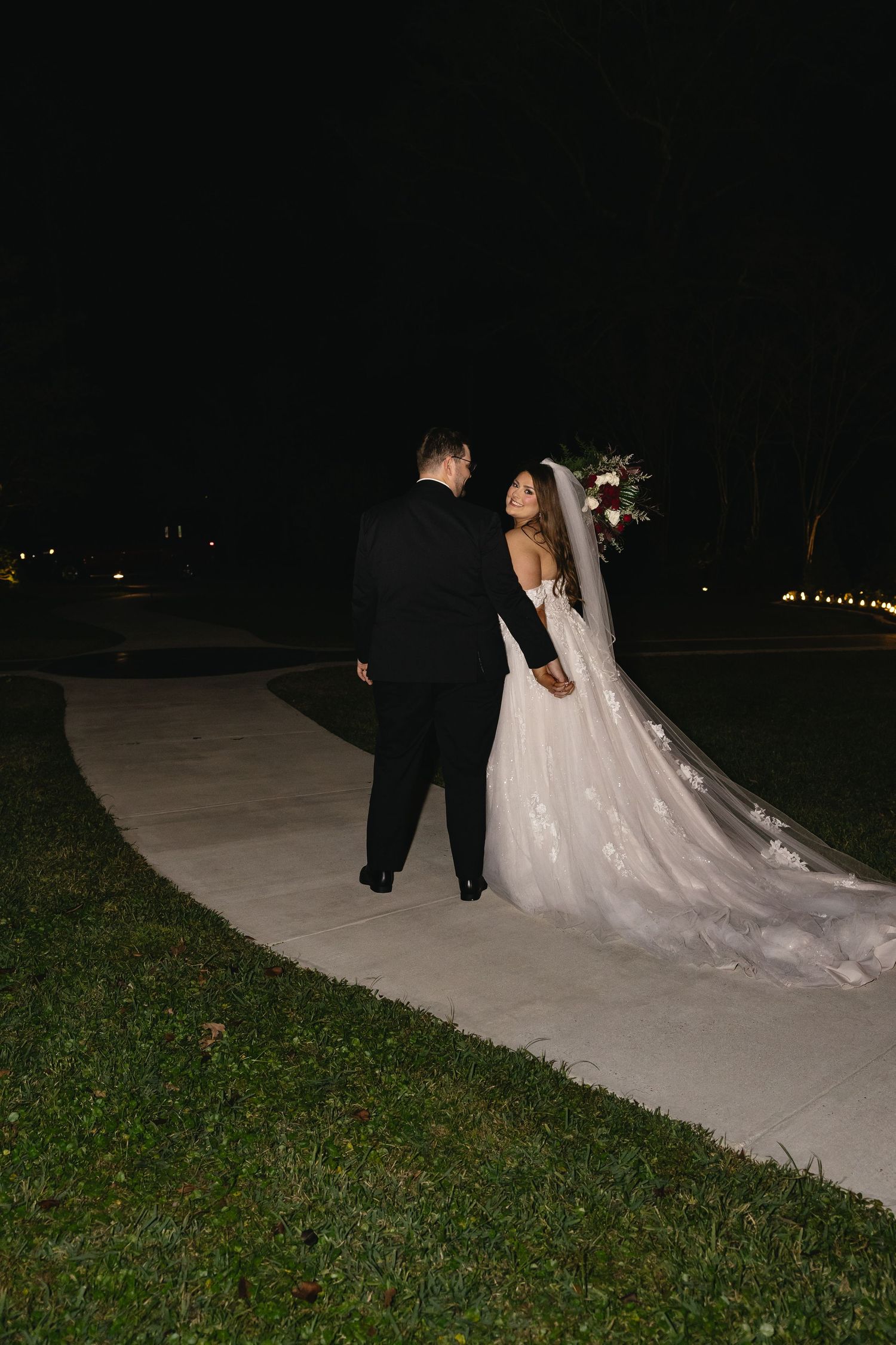 Newlyweds walk hand in hand along a lit pathway at night after their wedding ceremony.