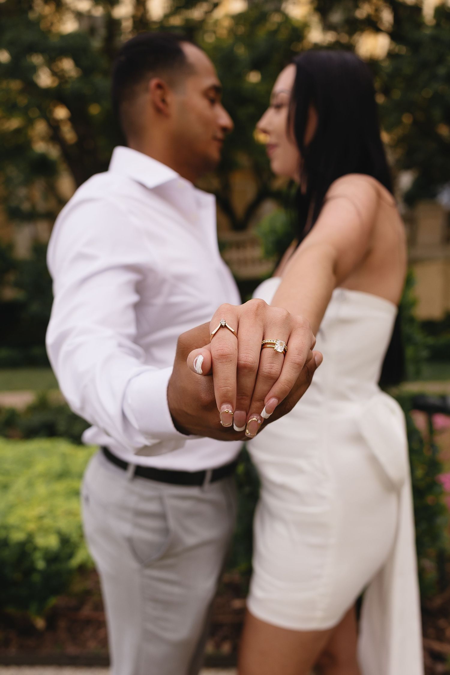 Engaged couple showing off wedding rings while holding hands outdoors, focus on gold engagement and wedding bands.