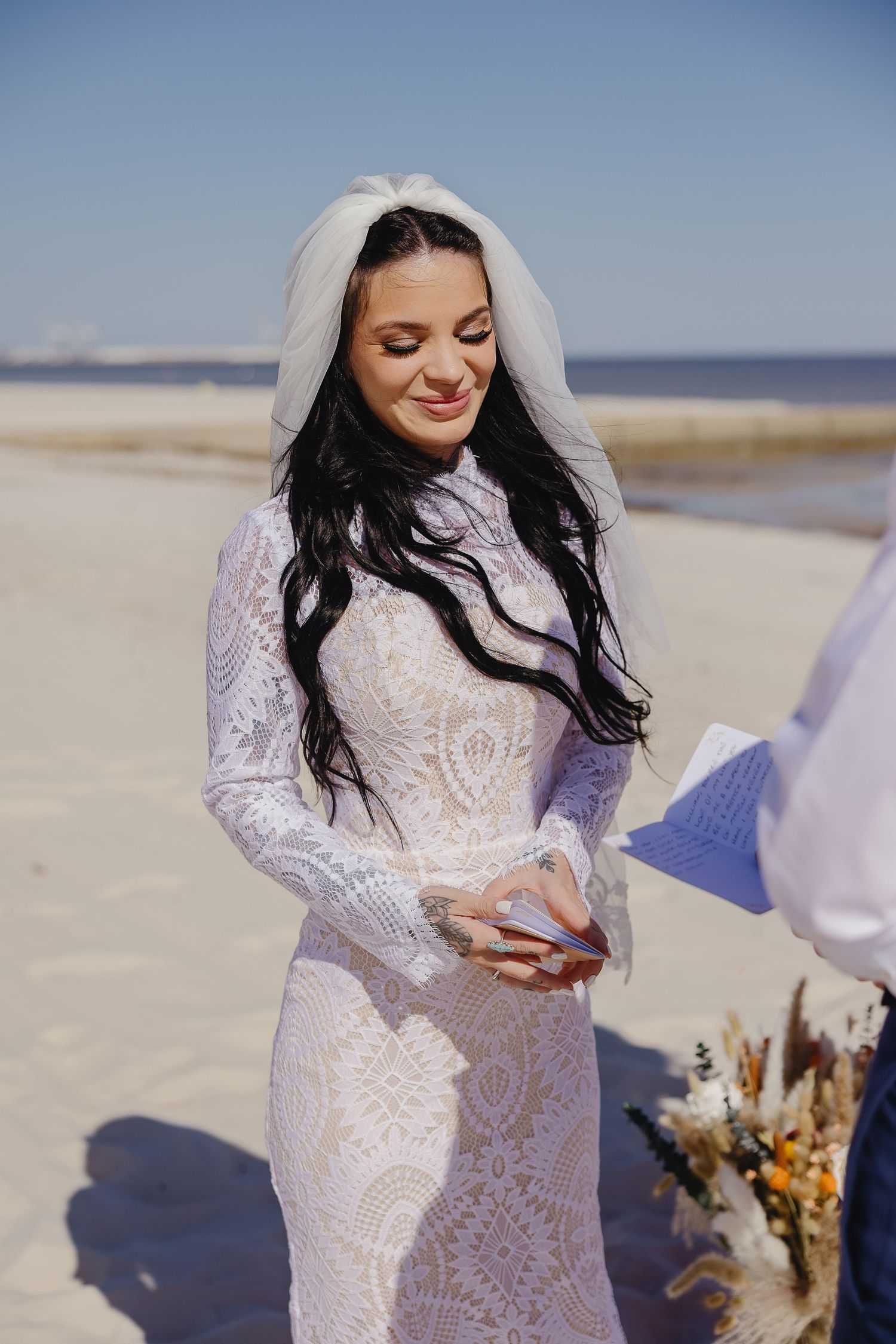 Dark-haired bride in long-sleeve lace wedding gown and veil smiling during beach wedding ceremony on sunny day.