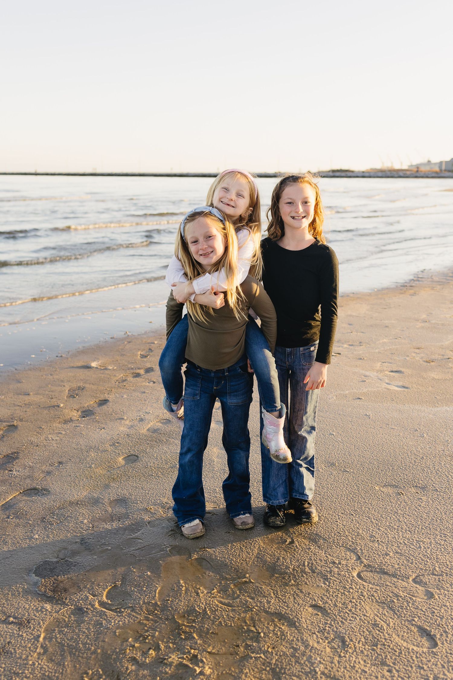 Three young girls smile and play piggyback on a sandy beach with ocean waves behind them.