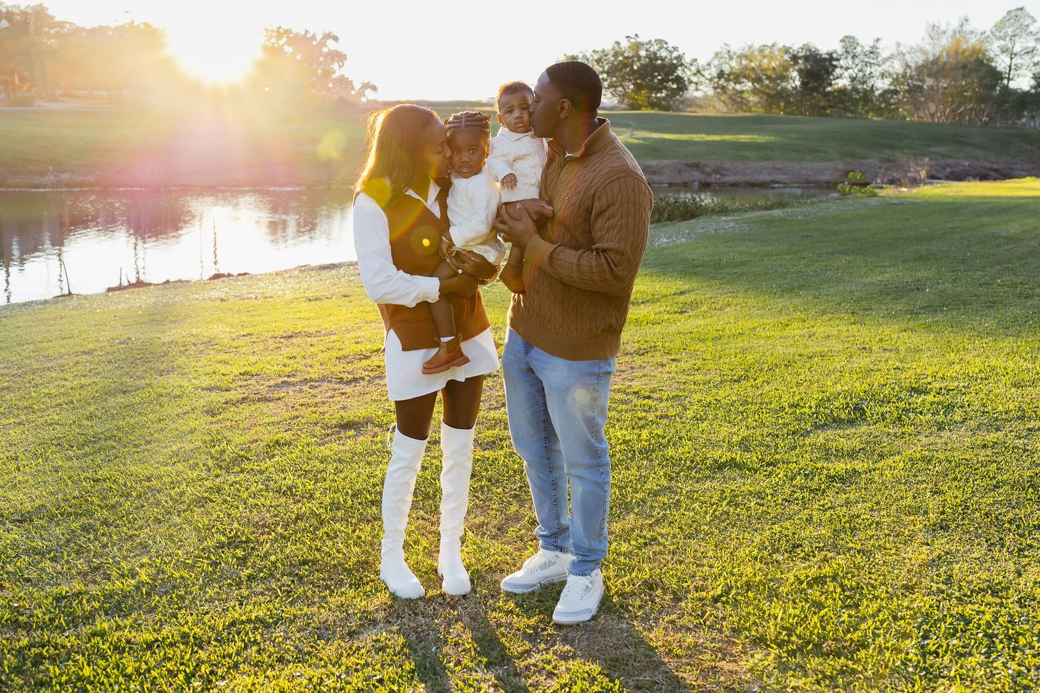 A family of three standing on green grass near a pond during a golden sunset.