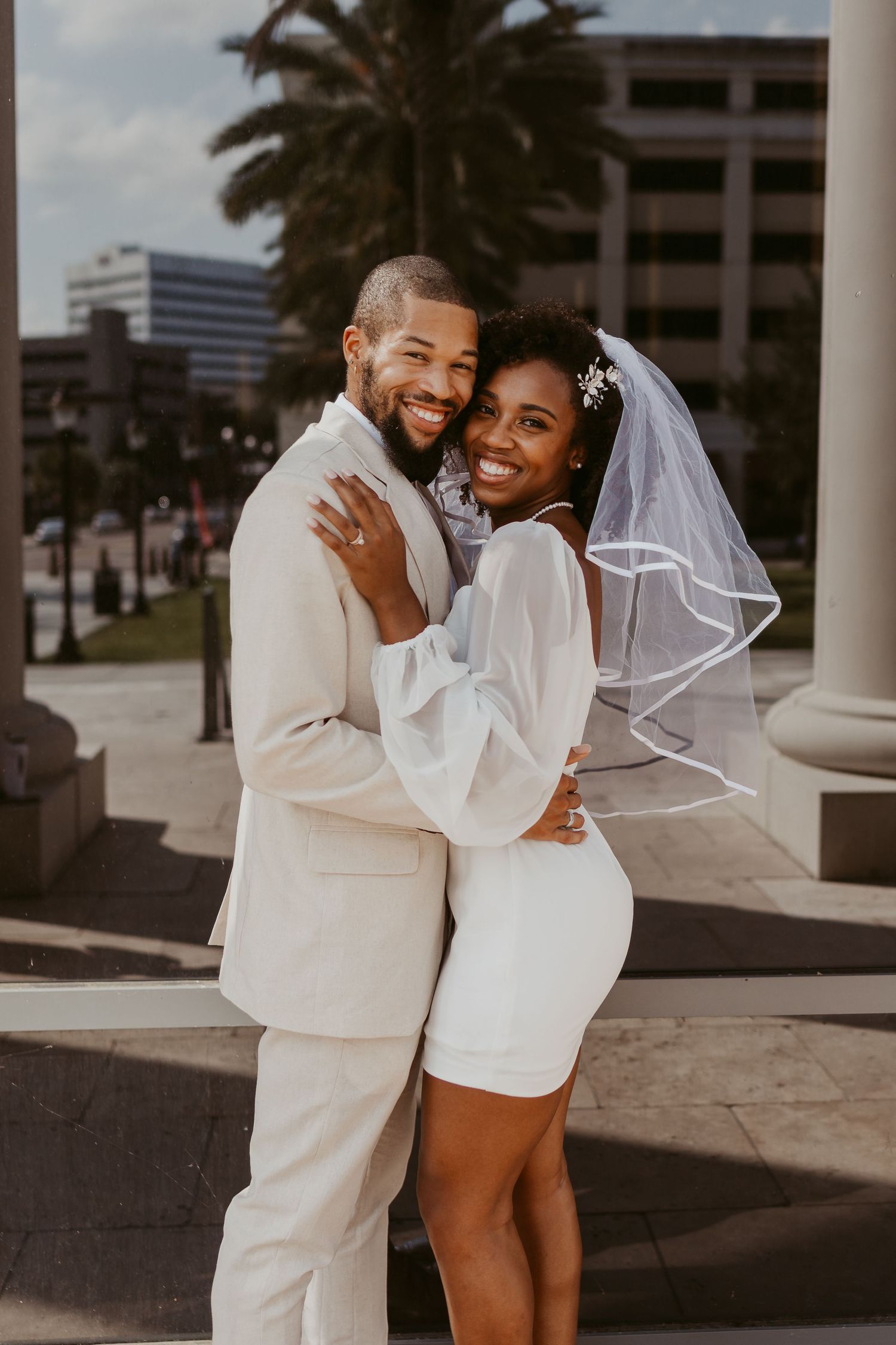 Smiling bride in short white dress and veil embracing her groom in a cream suit outdoors.