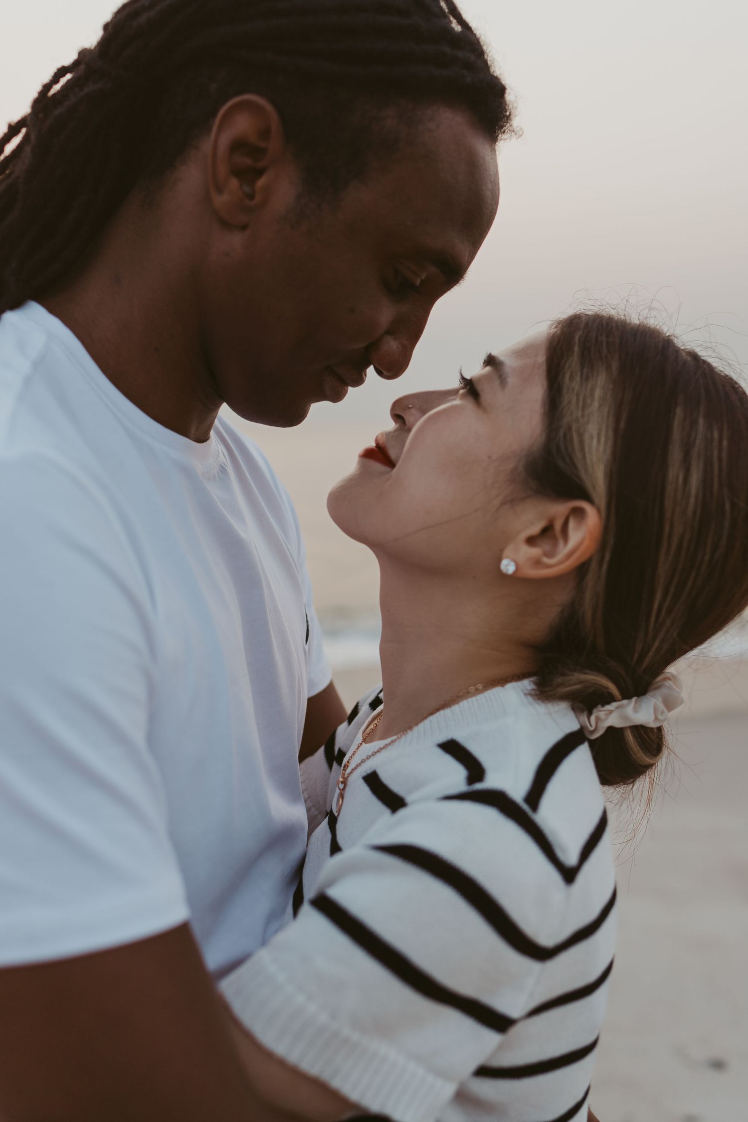 Interracial couple sharing an intimate moment on the beach at sunset, nearly kissing.