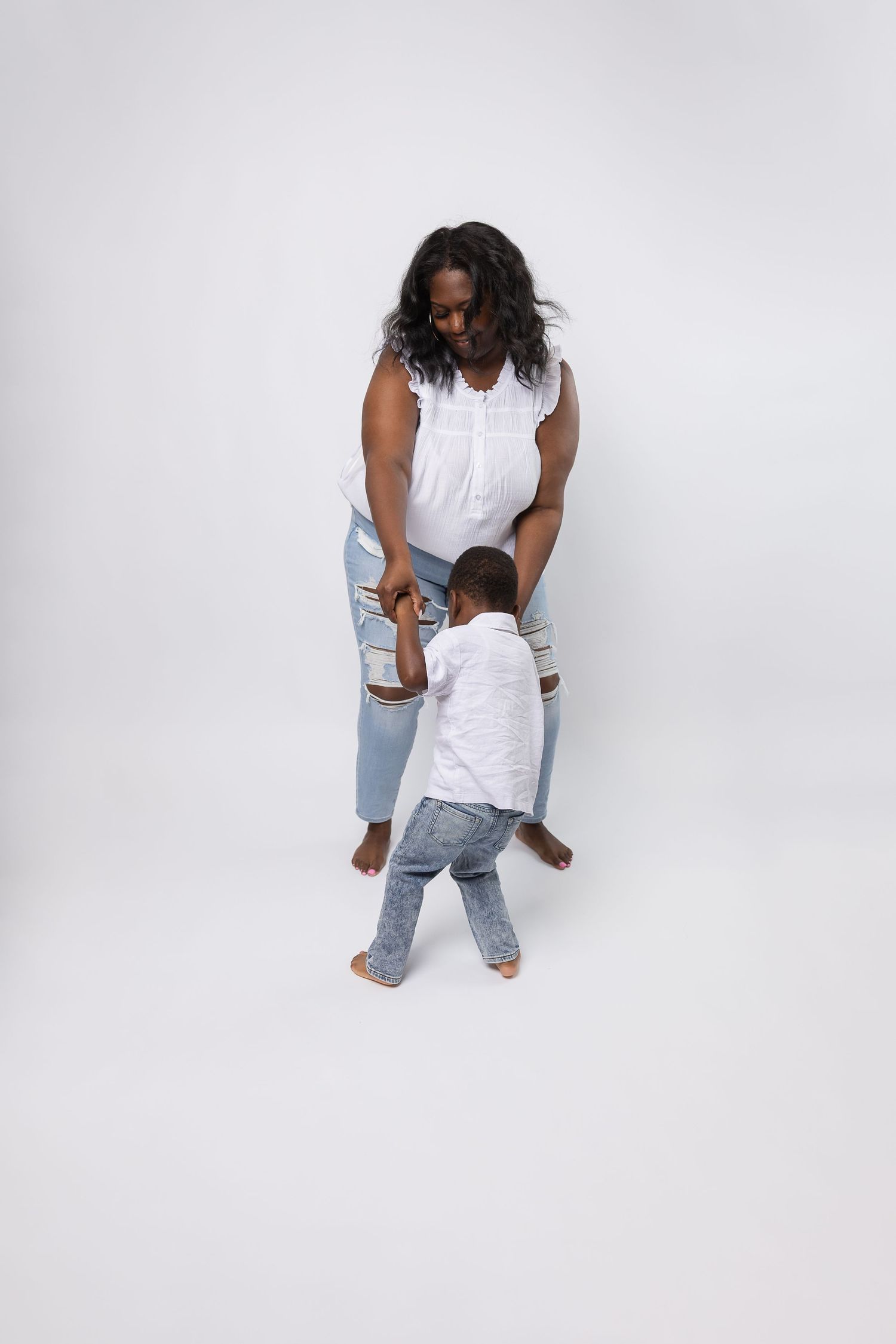 Mother in white top and jeans holds hands with toddler in white shirt on white background.