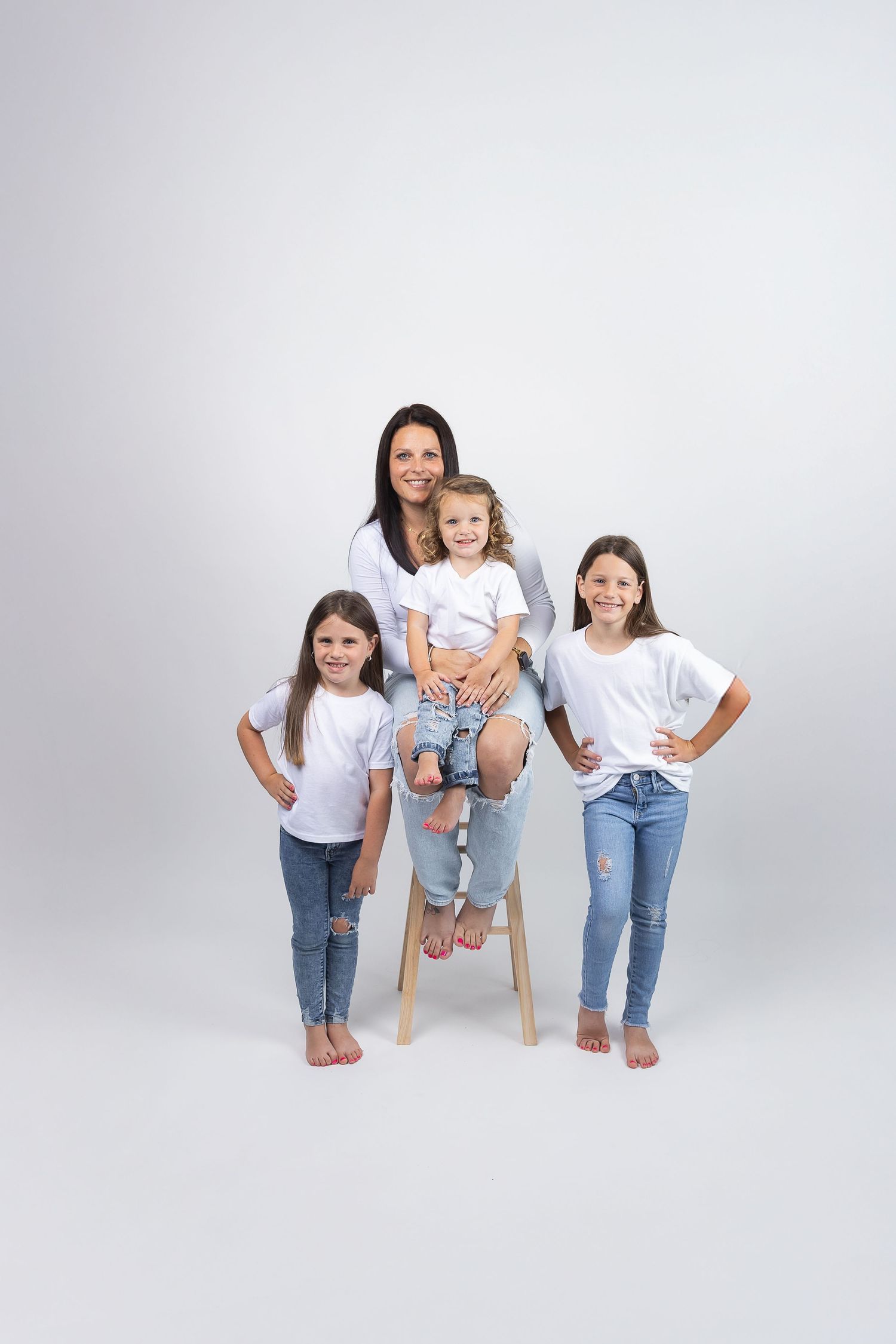 Mother and three daughters in matching white shirts and jeans pose together on white background.