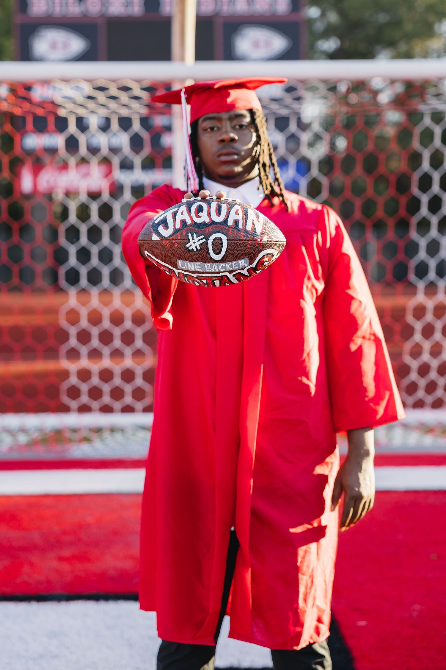 High school senior in red graduation gown holding custom football with name Jaquan #0 on field.