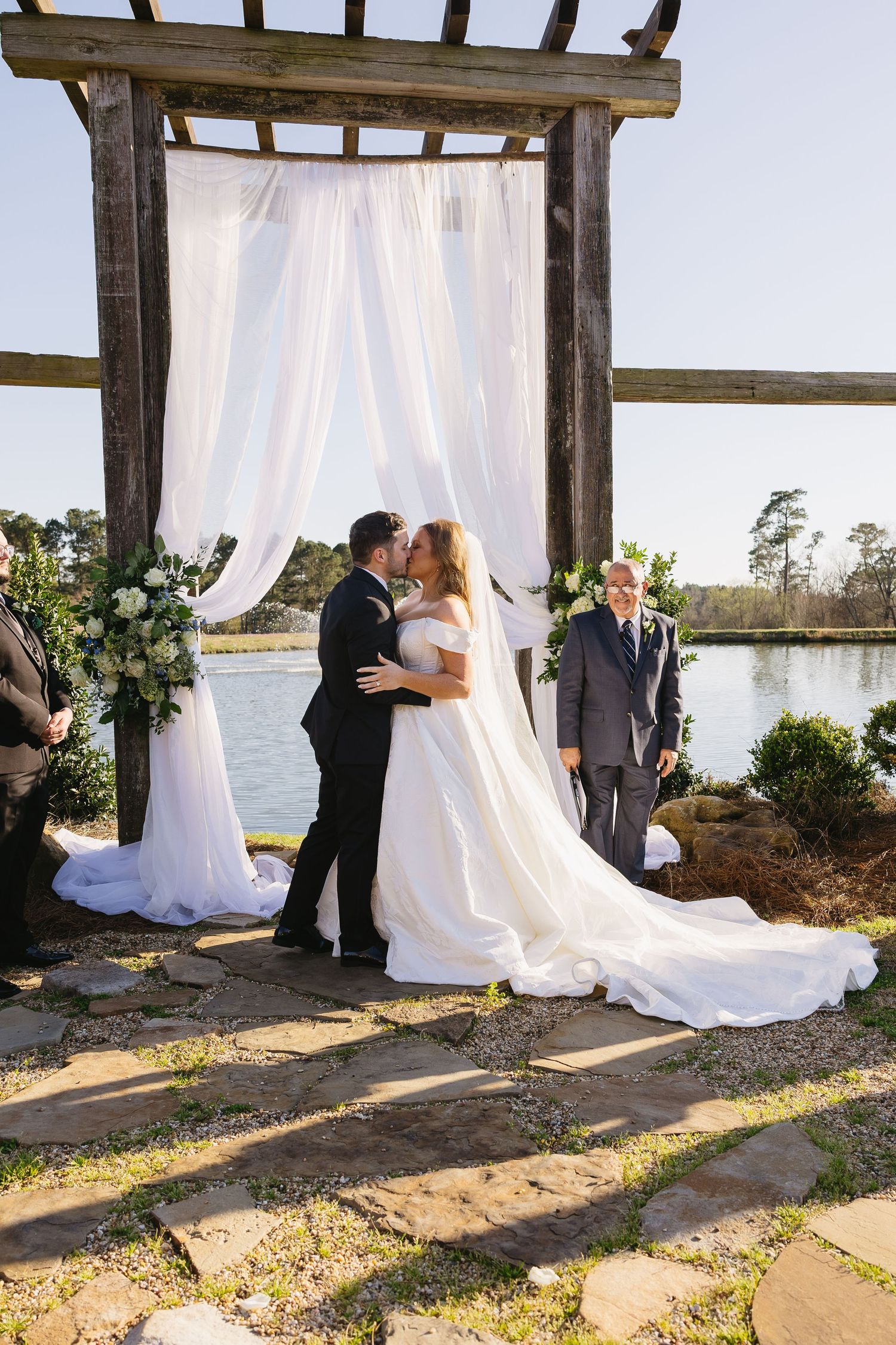 Bride and groom share first kiss at outdoor lakeside ceremony under white draped wooden arch.