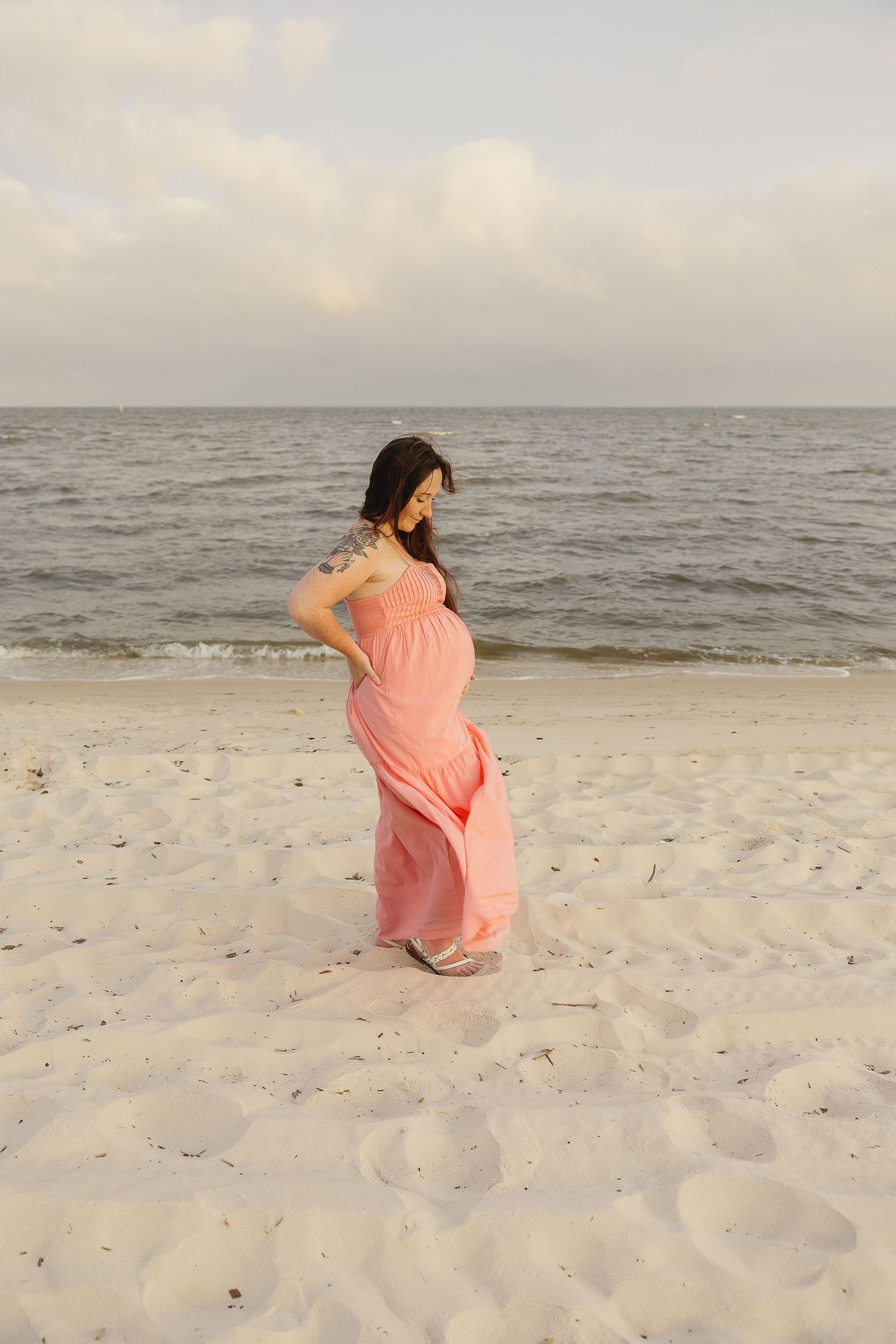 Pregnant woman in flowing pink dress posing solo on beach, wind blowing dress near ocean.
