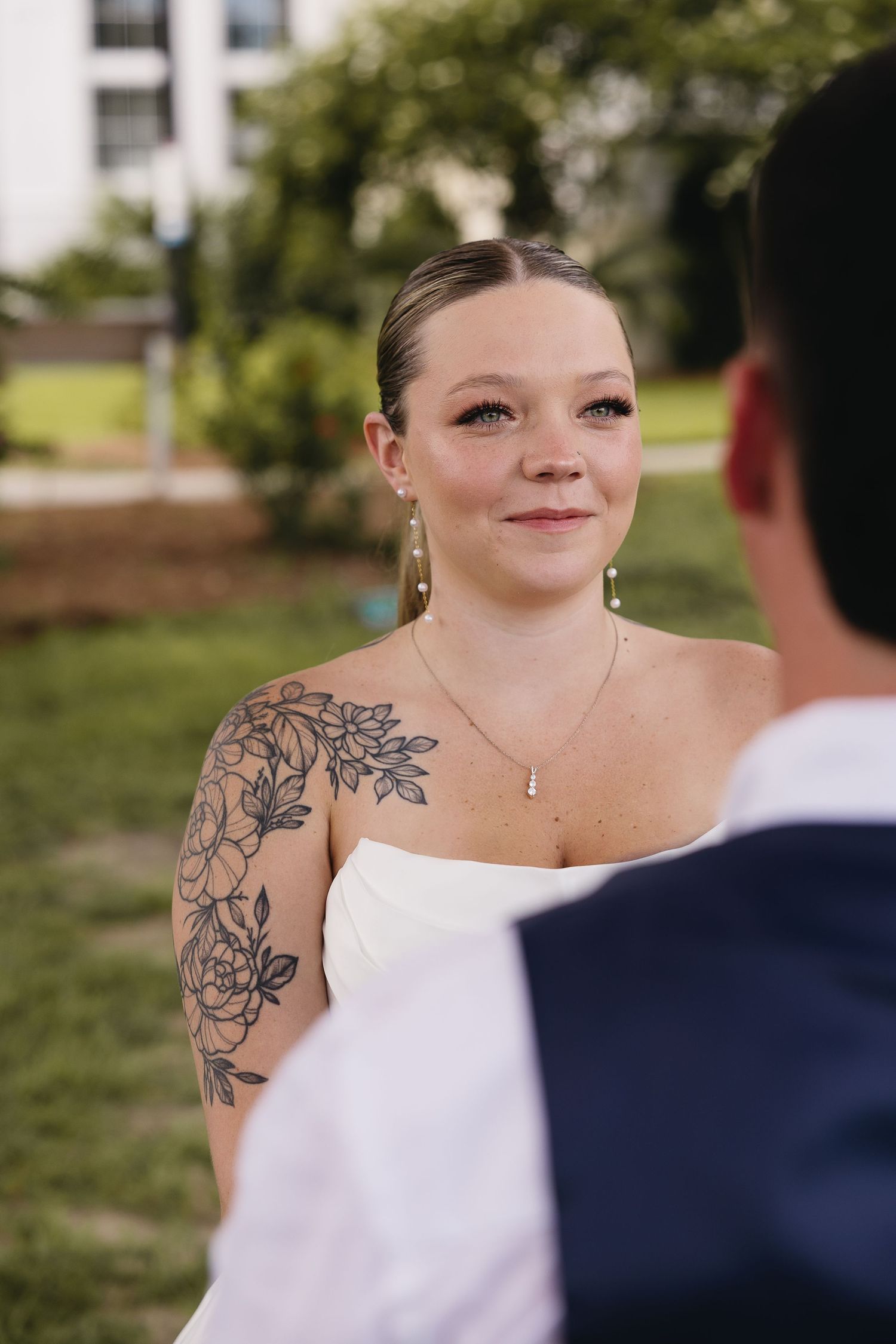 Bride with floral arm tattoo smiling during outdoor wedding ceremony.