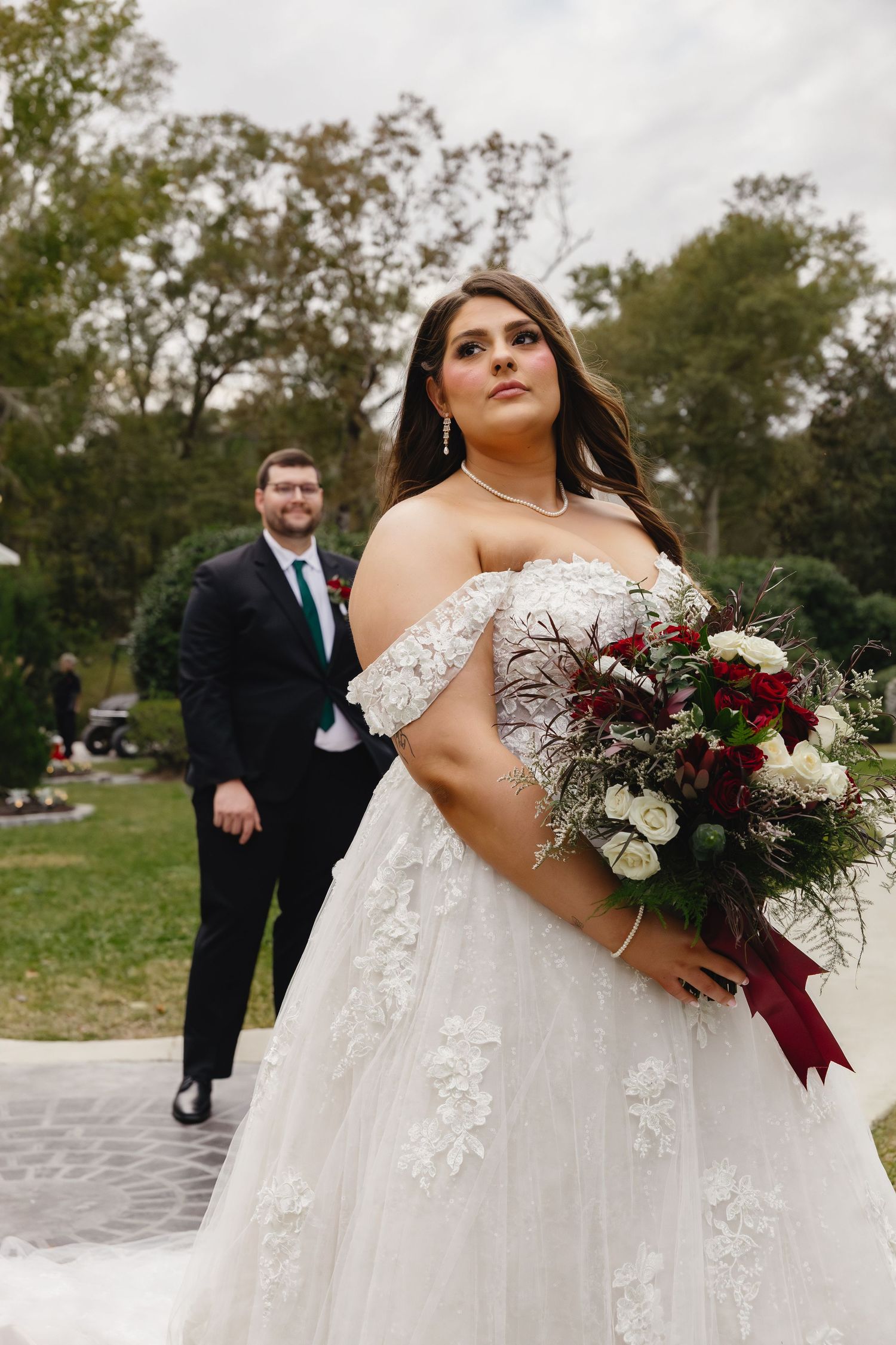 Bride in off-shoulder lace gown holds red bouquet while groom smiles in background outdoors.