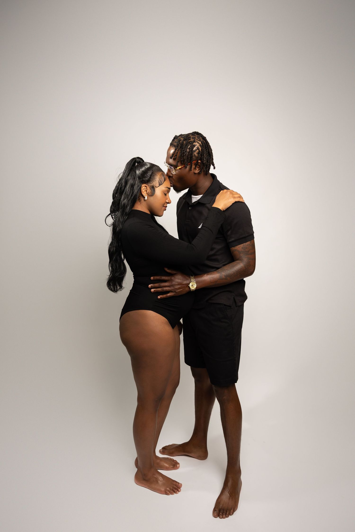 Couple in matching black outfits embrace intimately, faces close together on white backdrop.