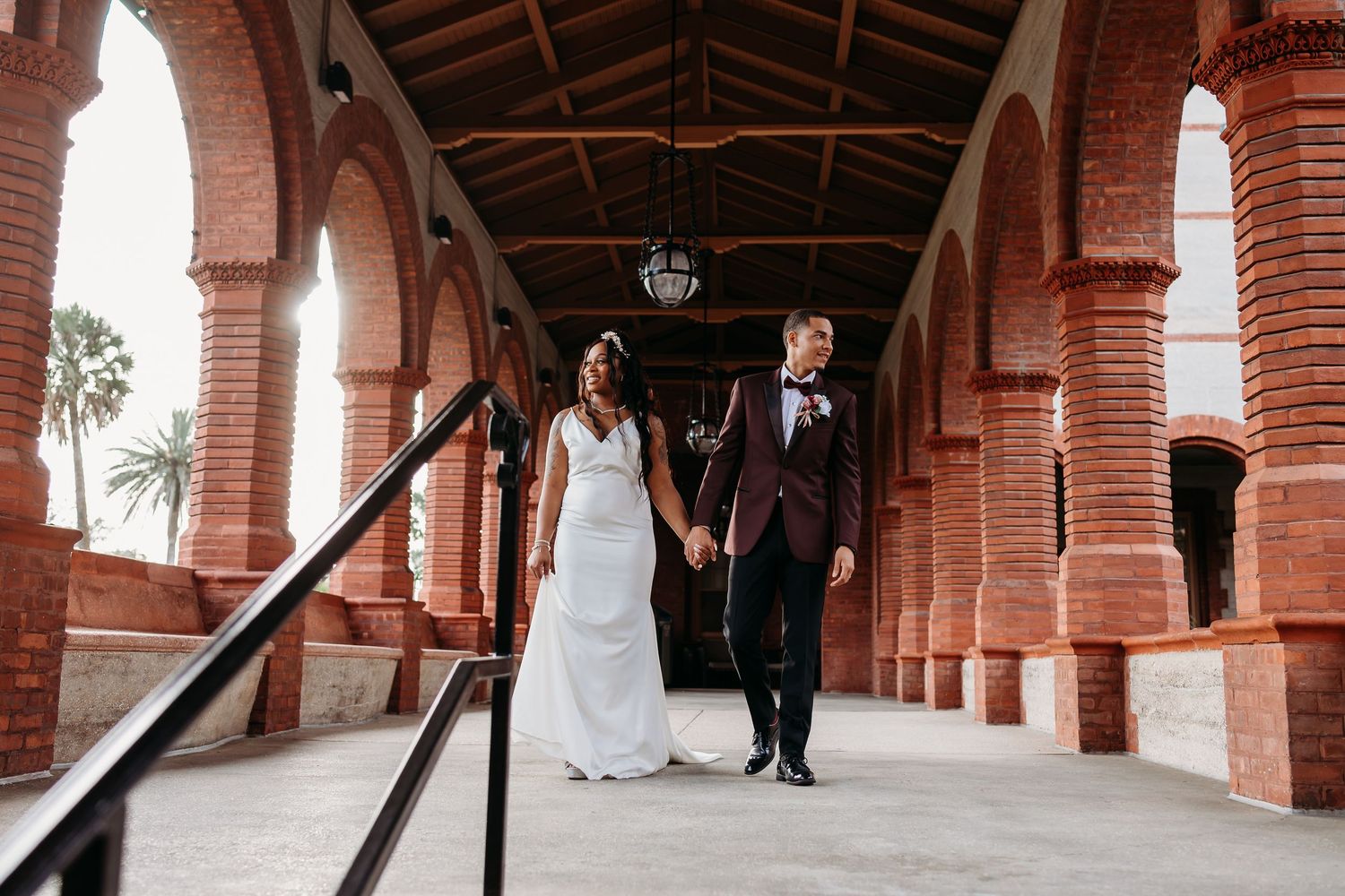 Bride and groom holding hands walking through a red brick arched corridor.
