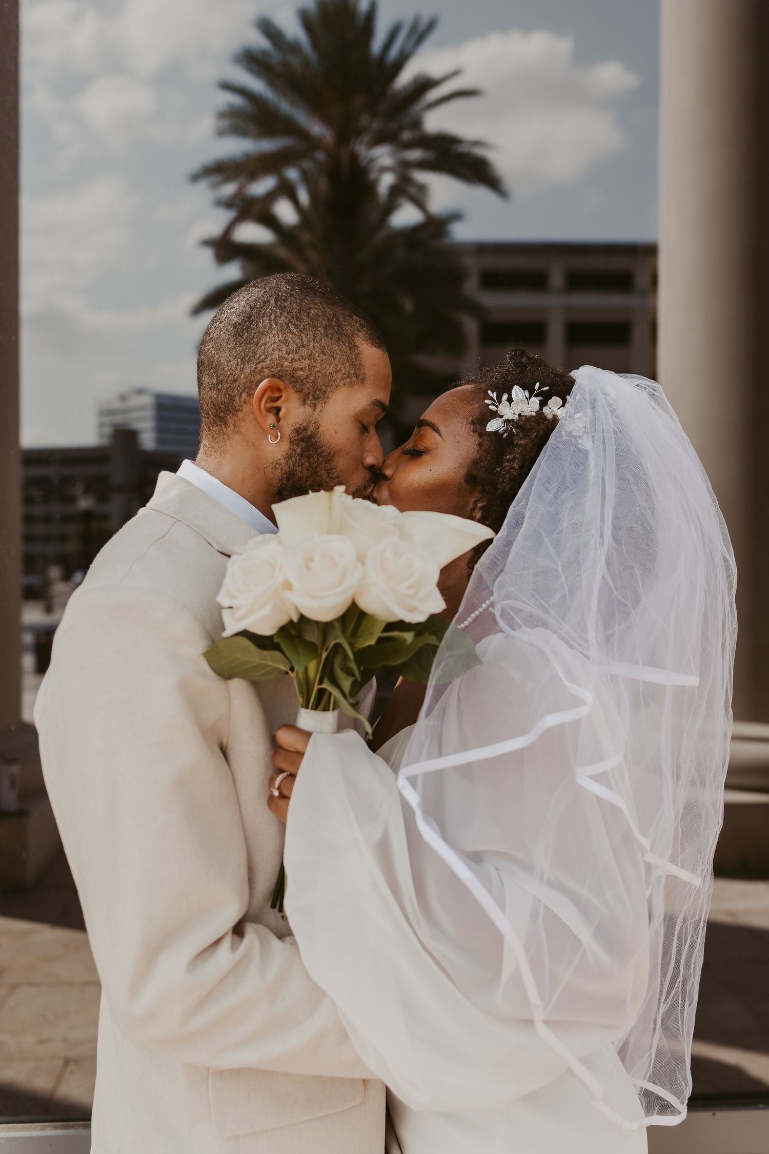 Newlywed couple sharing a kiss, bride holding white roses bouquet and wearing a flowing veil outdoors.