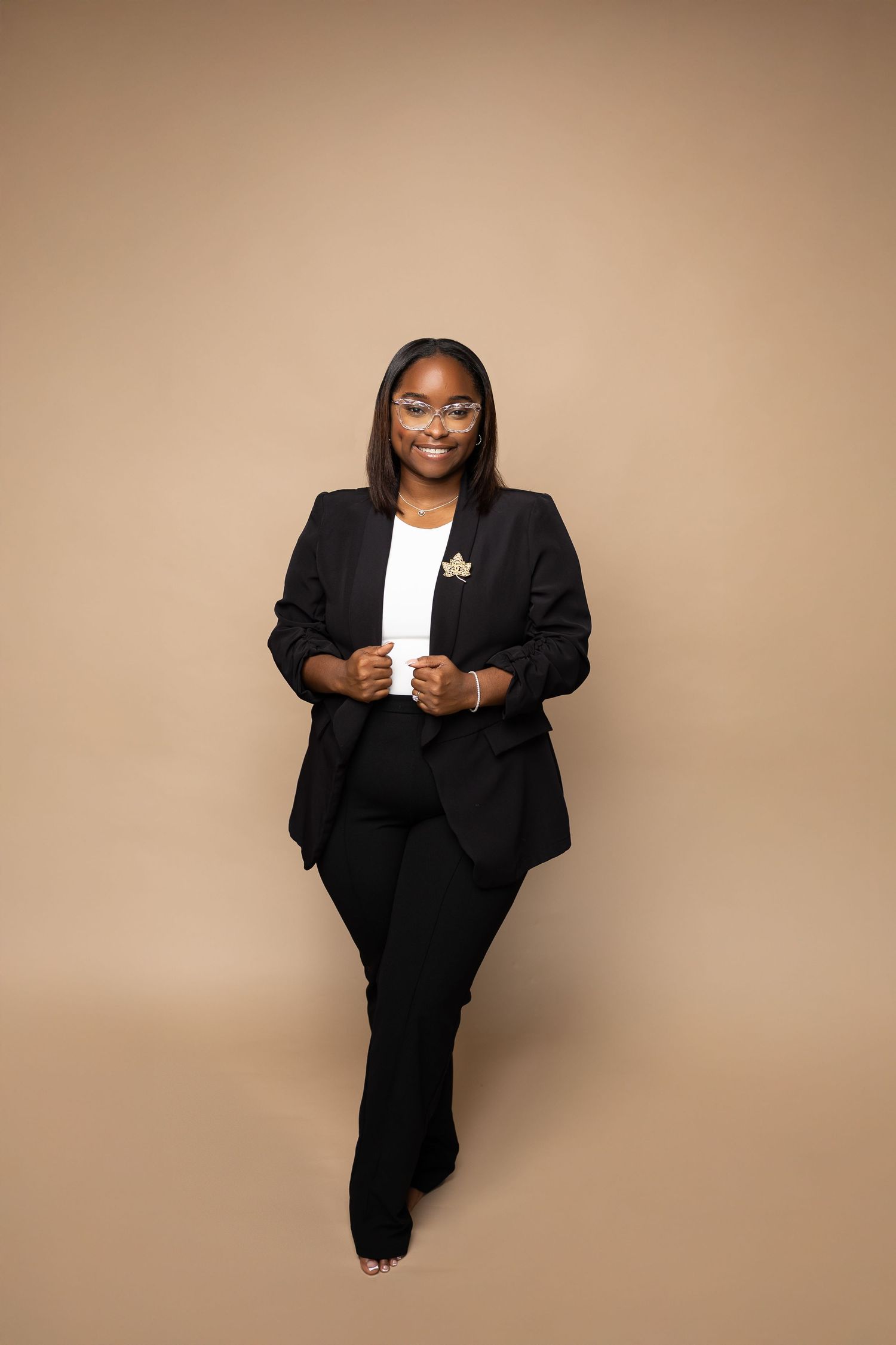 Smiling woman in black blazer and pants poses standing on beige studio background.