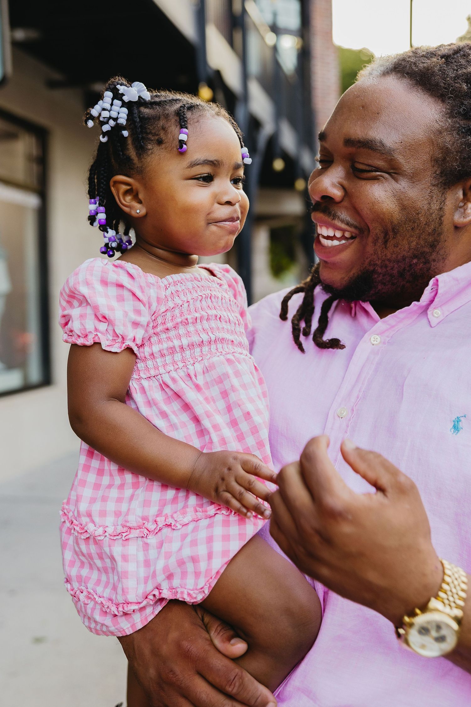 Laughing father holds smiling toddler daughter in pink gingham dress outdoors.