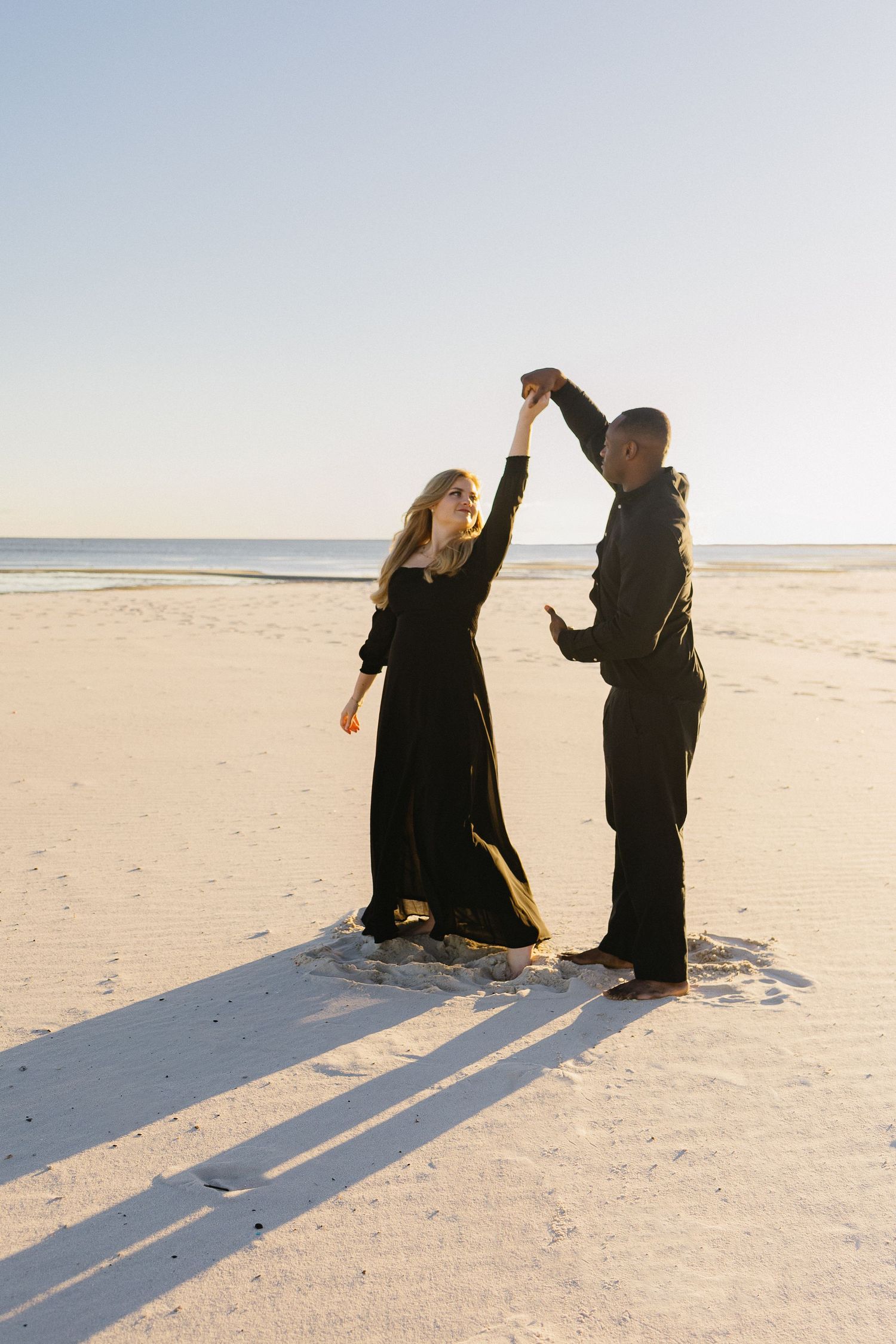 Couple dancing on a sandy beach at sunset, both dressed in elegant black outfits, holding hands raised.