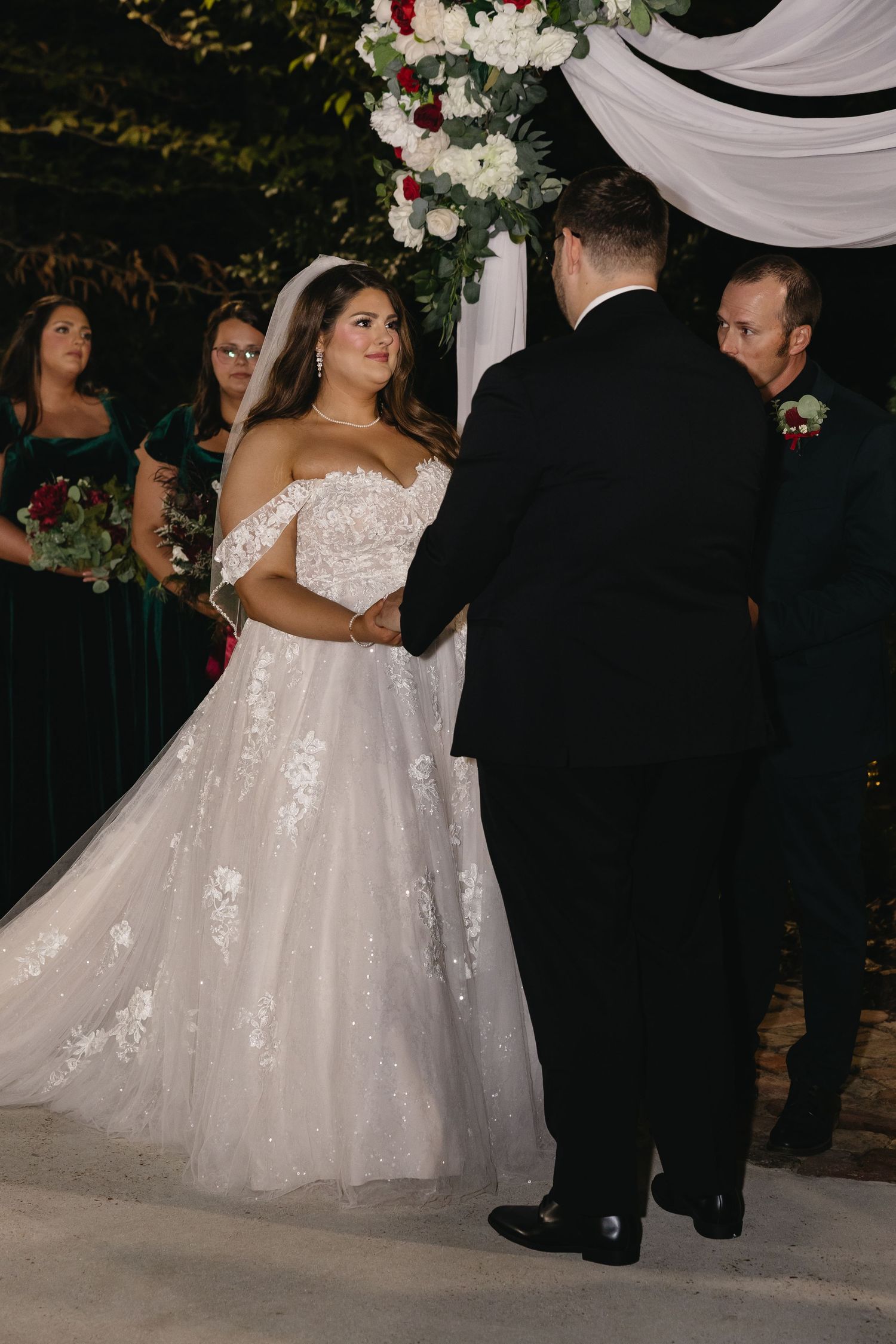 Bride in lace ball gown holds groom's hands at outdoor altar.