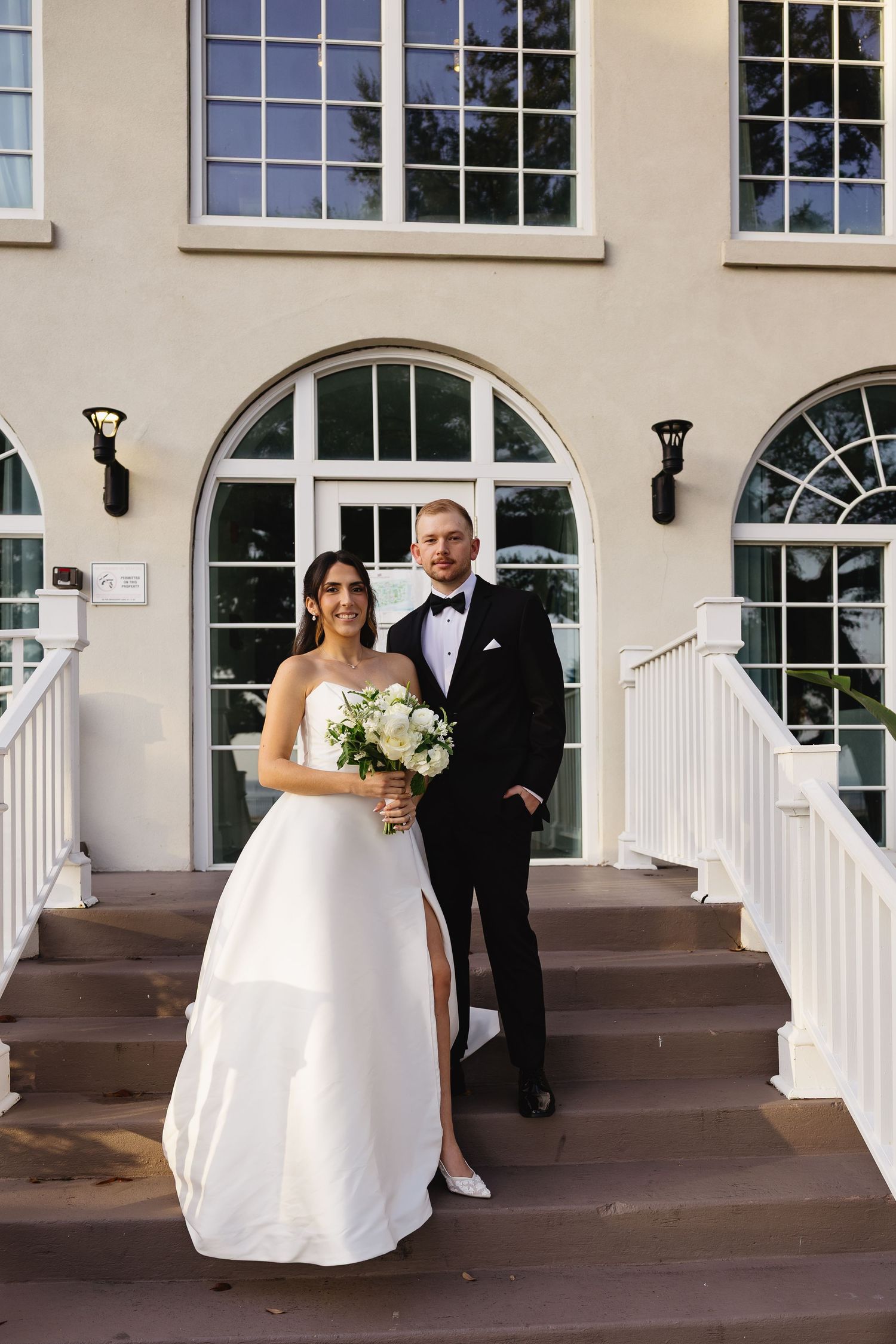Bride holding white floral bouquet and groom in tuxedo stand on steps outside an elegant venue.