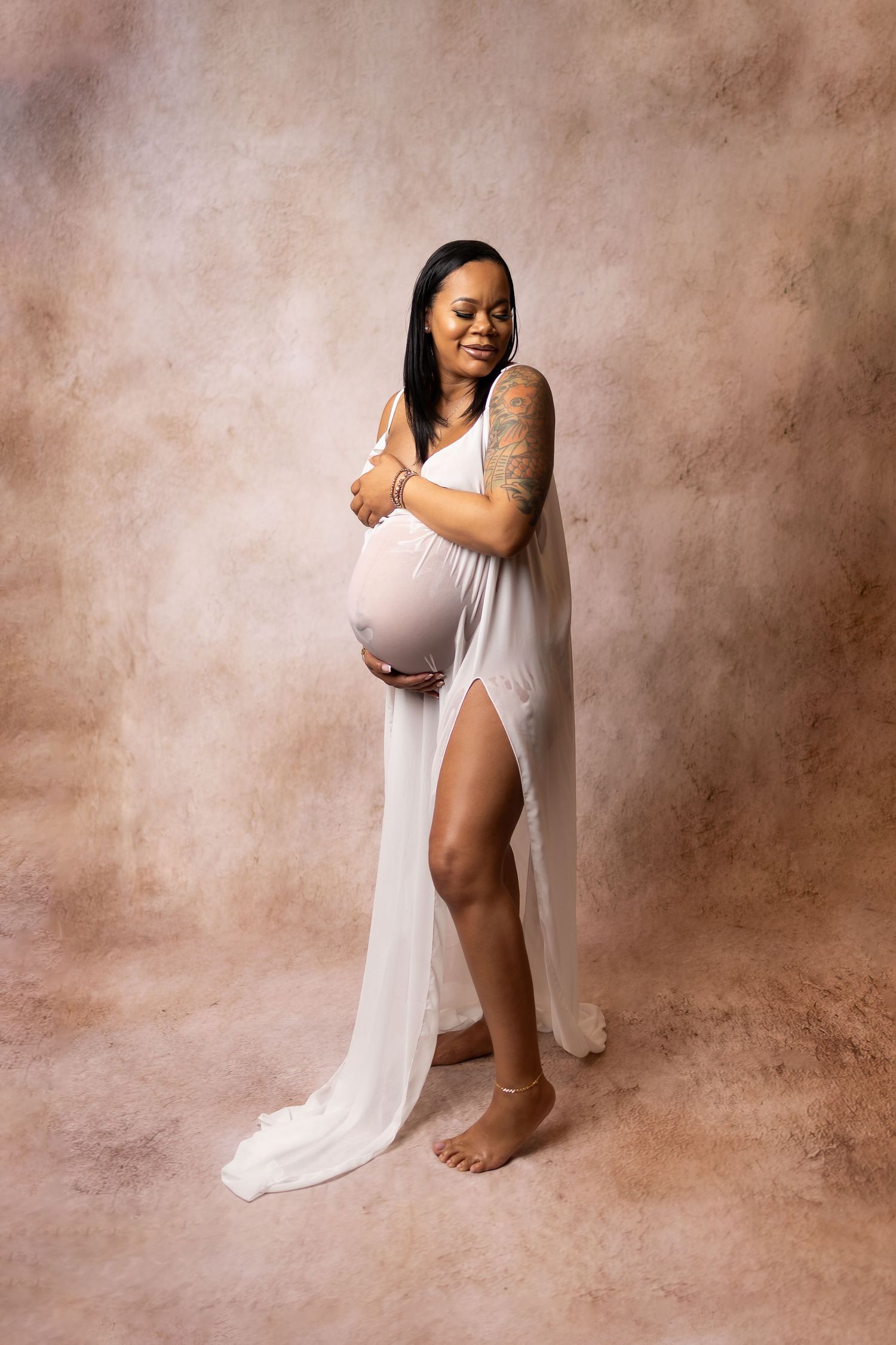 Pregnant Black woman in sheer white draped gown cradles baby bump on textured brown backdrop.
