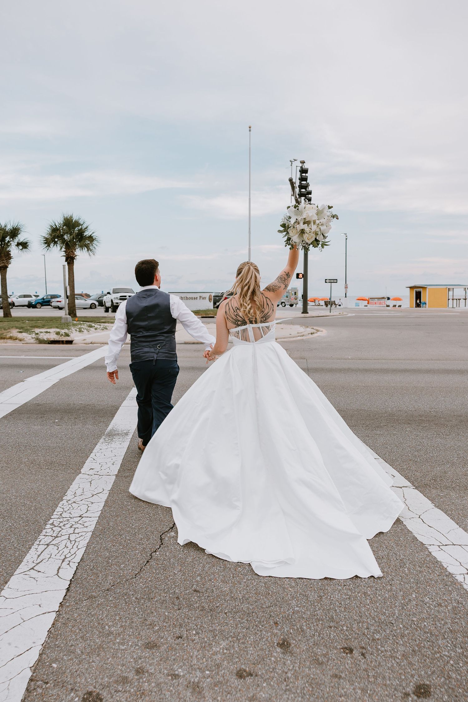 Bride raising white floral bouquet while walking with groom across a crosswalk near the waterfront.