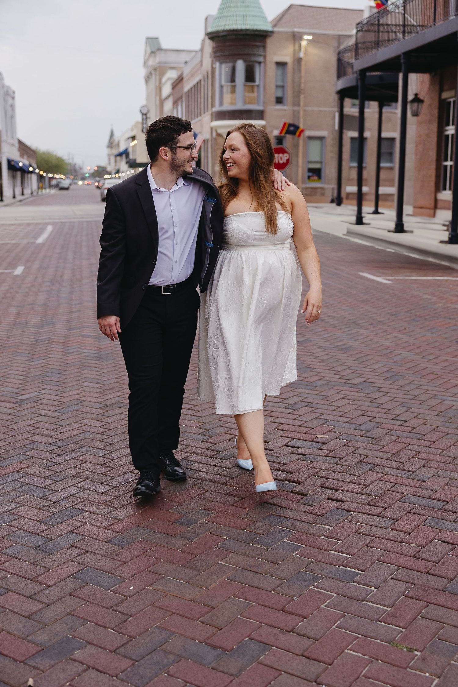 Happy couple walking together on brick street, woman in white midi dress with light blue heels and man in black suit.