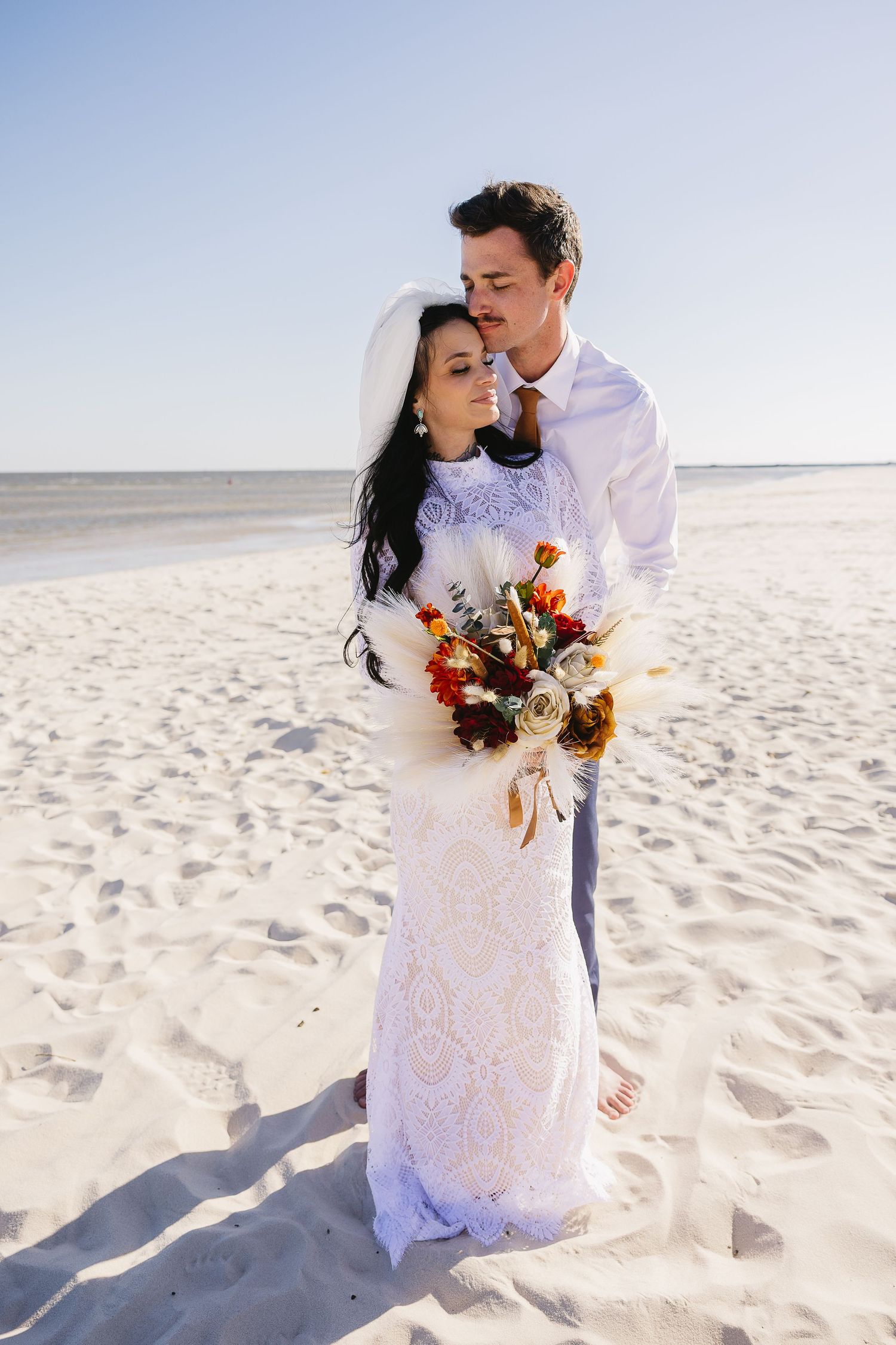 Bride and groom embrace on a sunny white sand beach, bride holding colorful bouquet.