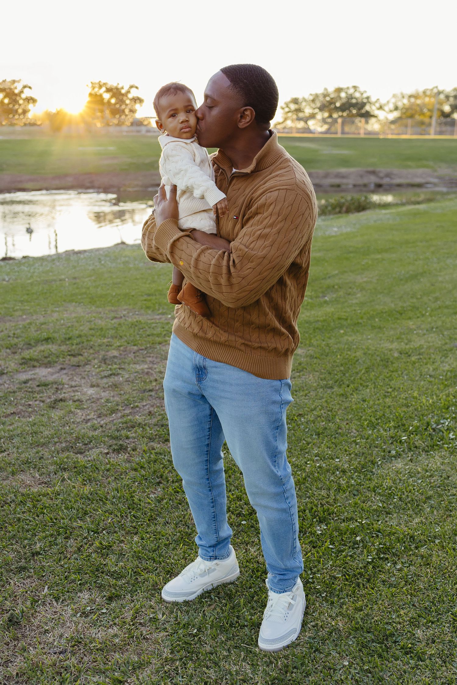 Loving Black father kissing baby on forehead while standing on grass near a pond at sunset.
