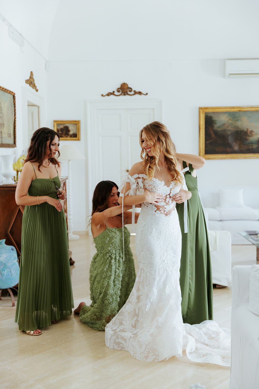 Group of bridesmaids in green dresses helping with white wedding gown preparations in a bright room.