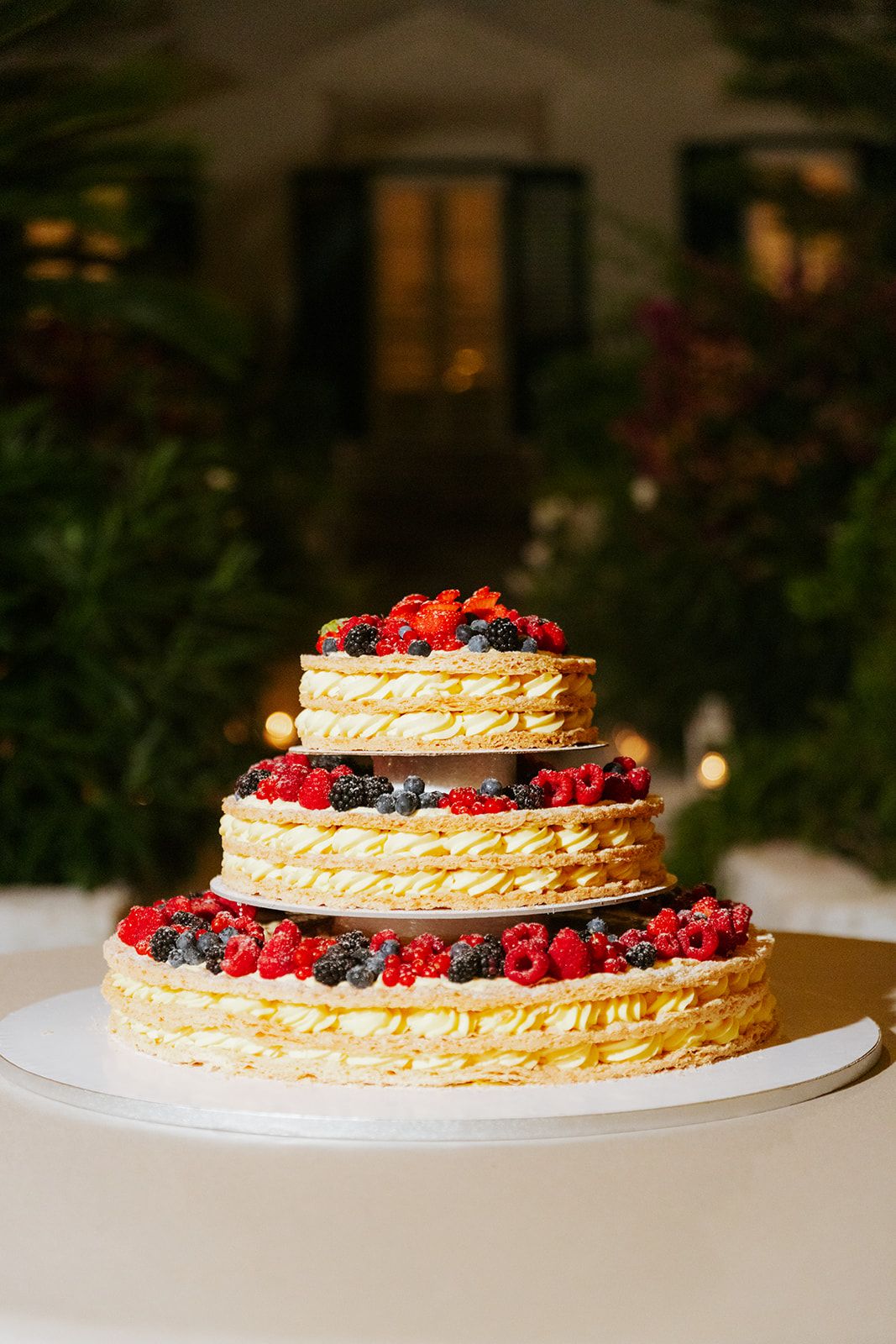 Three-tiered layered cake decorated with fresh red and blue berries on a white cake stand against a dark background.