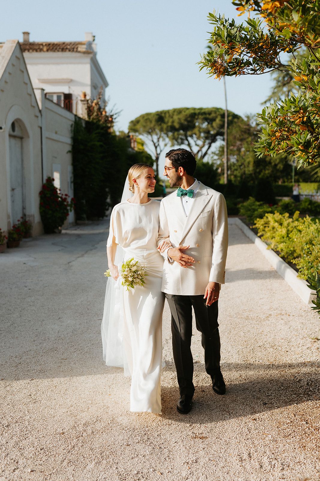 A wedding couple walks hand in hand along a sun-dappled garden path beside a white Mediterranean-style building.