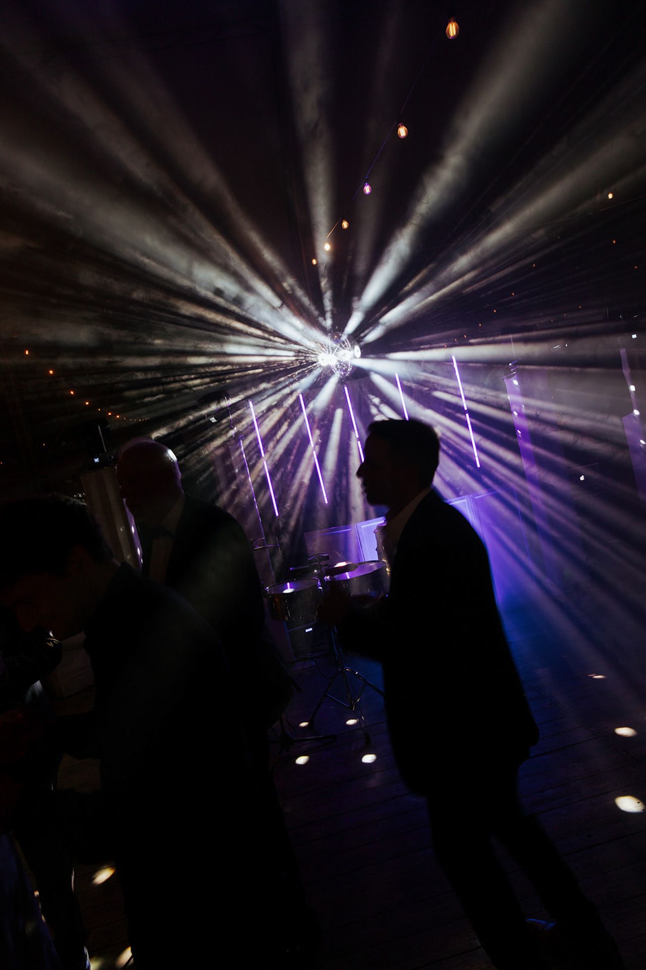 Silhouetted figures stand against dramatic purple and white stage lighting beams creating an atmospheric concert scene.