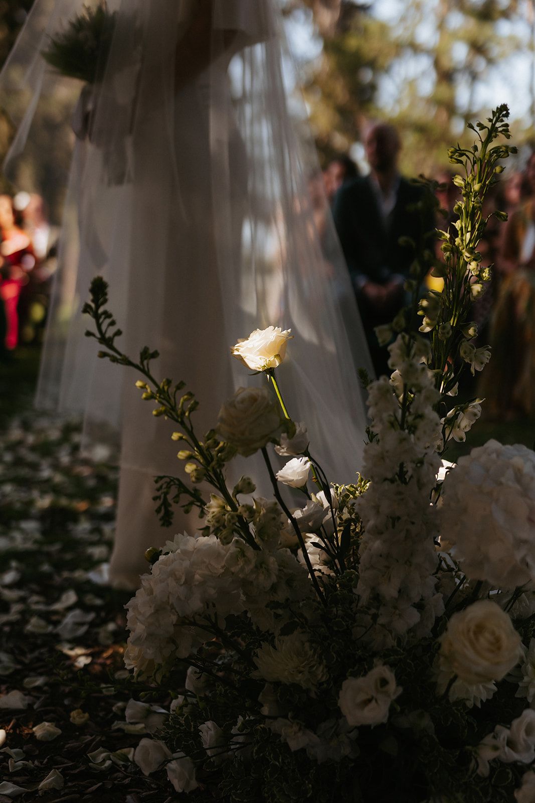 White roses and floral arrangements illuminate the bottom of a wedding dress veil in soft sunlight during an outdoor ceremony.