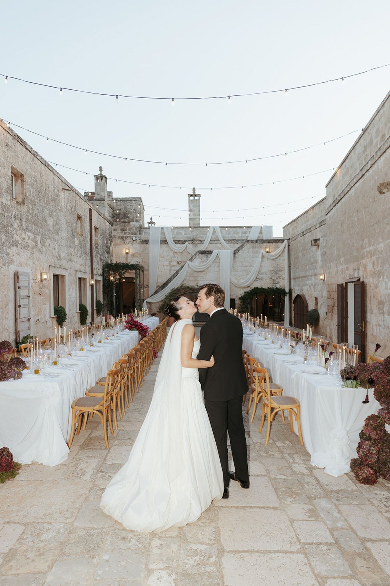 A wedding couple walks down a long stone path lined with wooden chairs and white linens in a rustic outdoor venue.