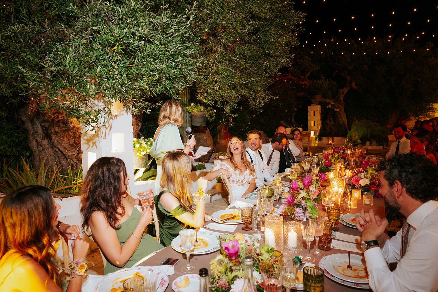 Wedding guests enjoy dinner at long tables decorated with floral arrangements under string lights in an outdoor garden setting.