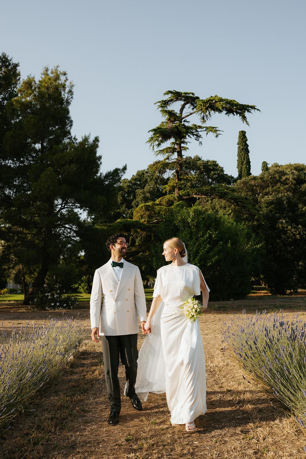Newlyweds walk hand in hand through a lavender garden path with cypress trees in the background at sunset.