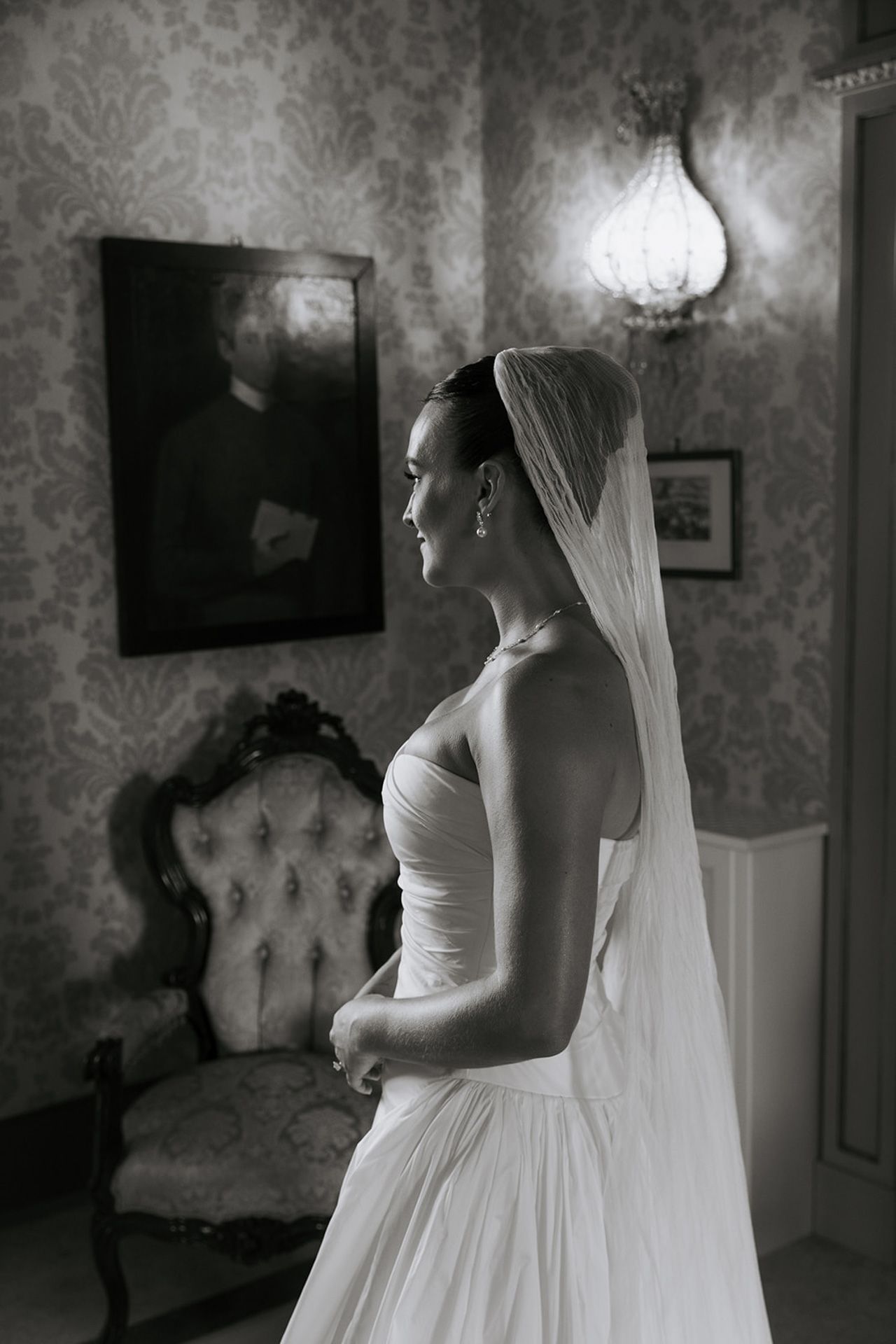 A bride in a classic white wedding gown stands in a vintage-style room with floral wallpaper and elegant lighting.