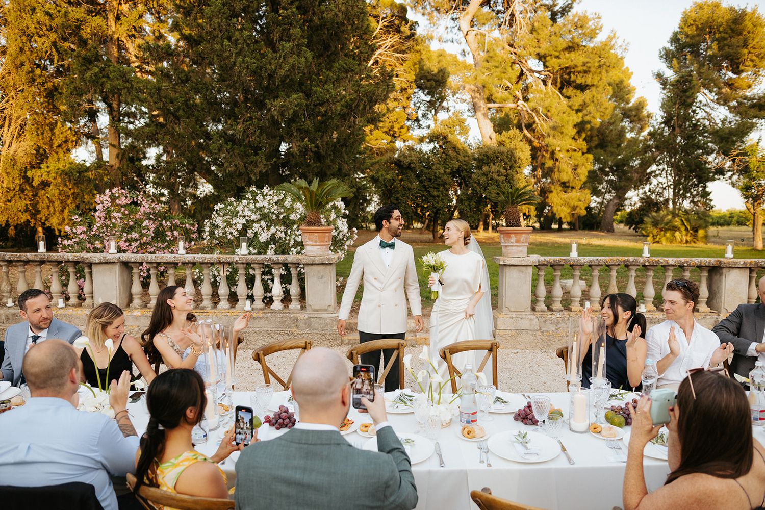 Elegant outdoor wedding reception with guests seated at tables under golden autumn trees near stone balustrade.
