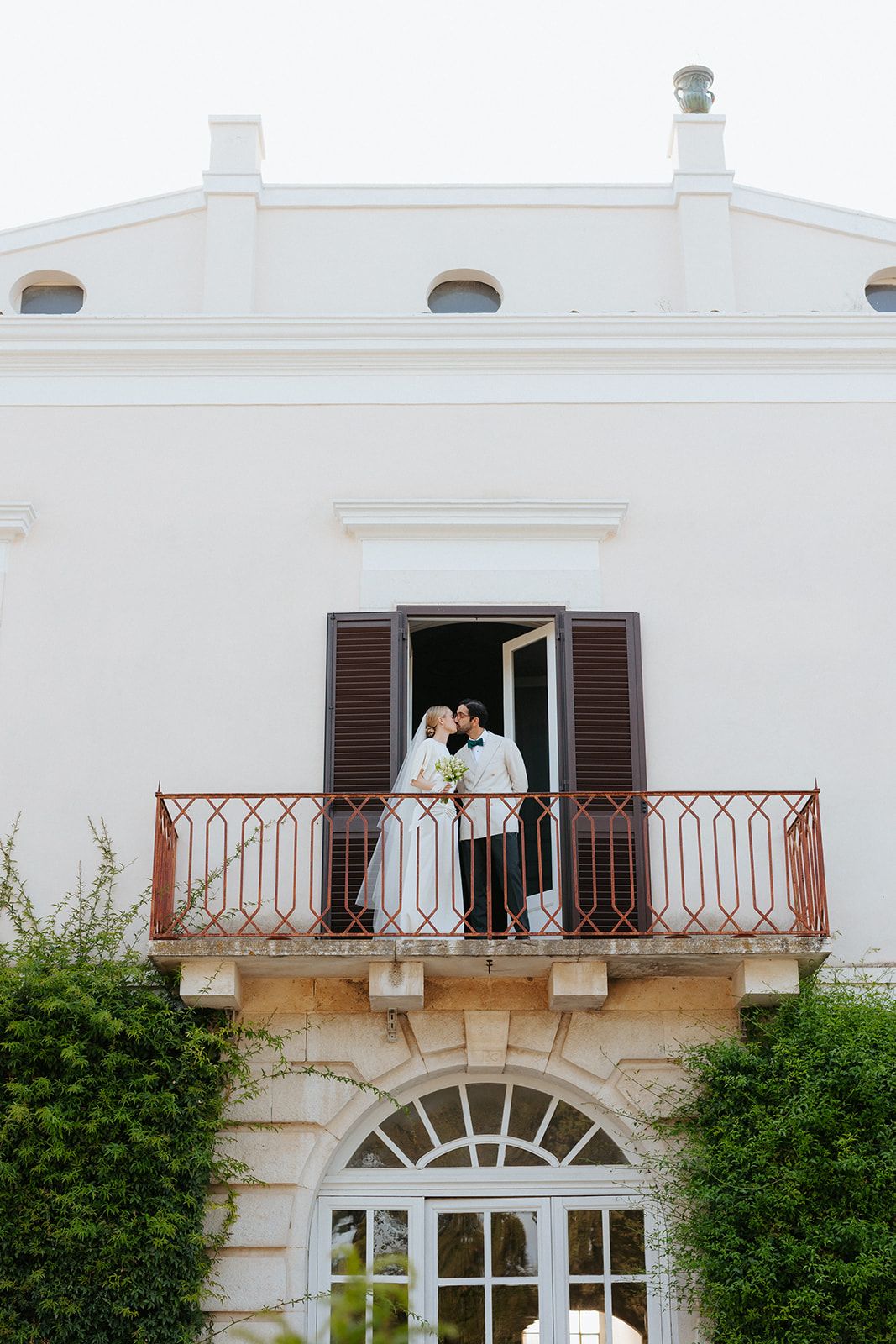 A white Mediterranean-style villa with brown shuttered windows and wrought iron balconies overlooking a lush garden.