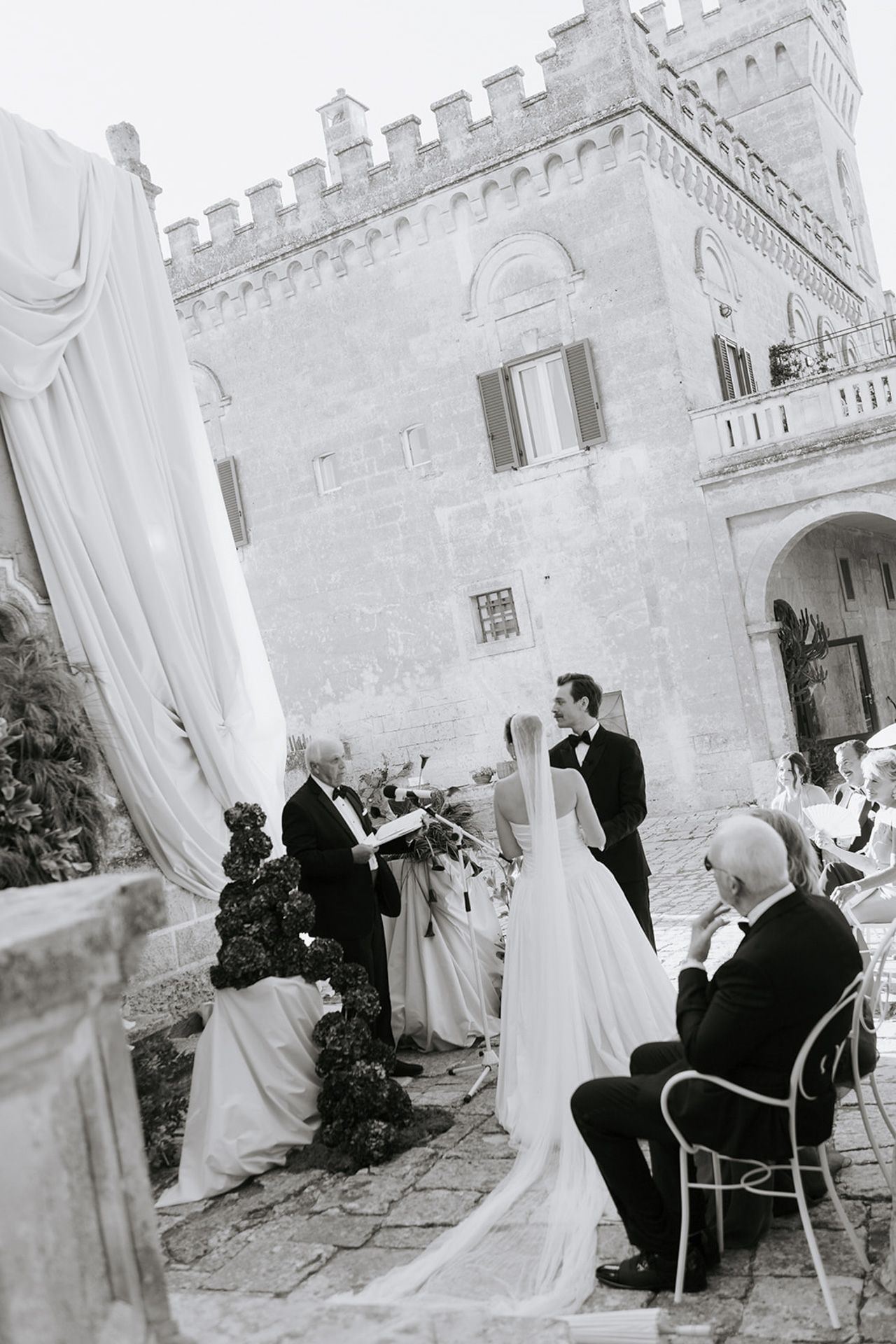 A sequence of black and white wedding photos showing guests seated outdoors at a castle-like venue with white drapes.