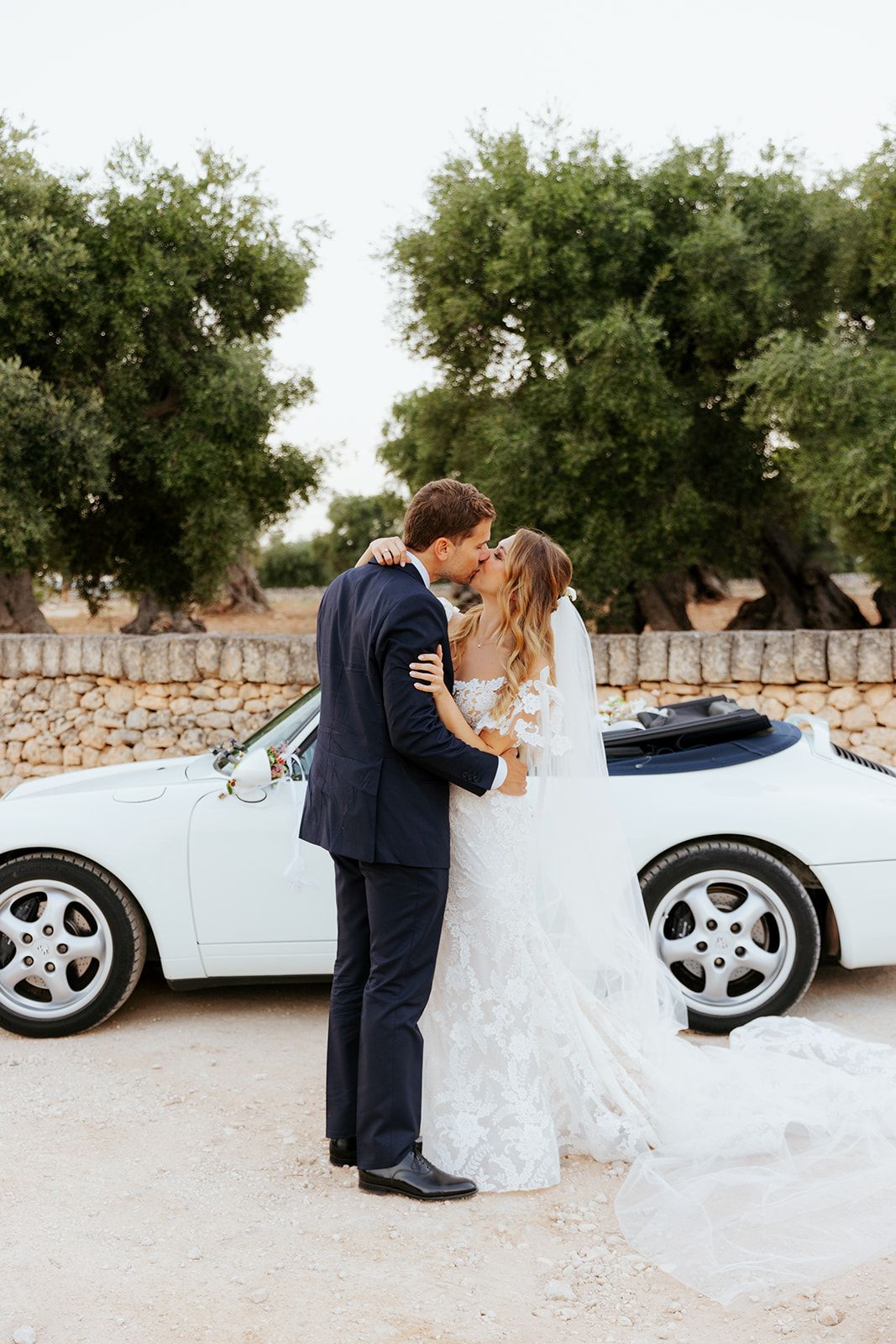 Newlyweds share intimate moments beside classic powder blue vintage car against stone wall backdrop.