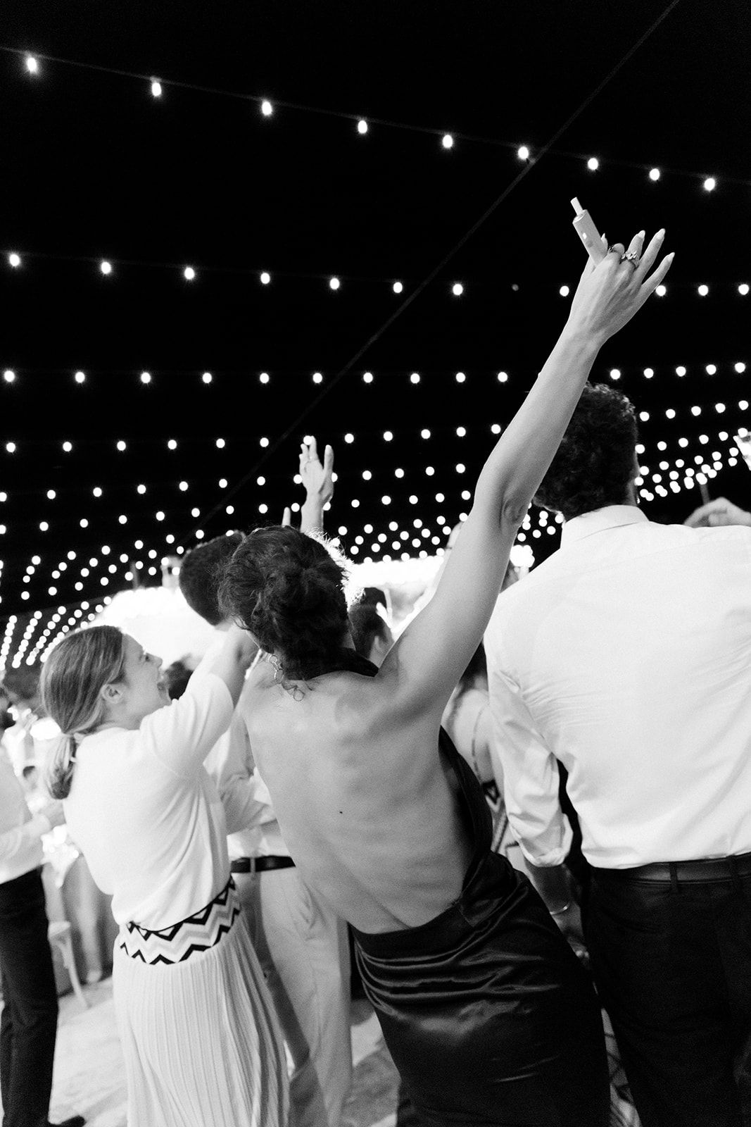 People with raised arms dancing at a celebration under decorative string lights.