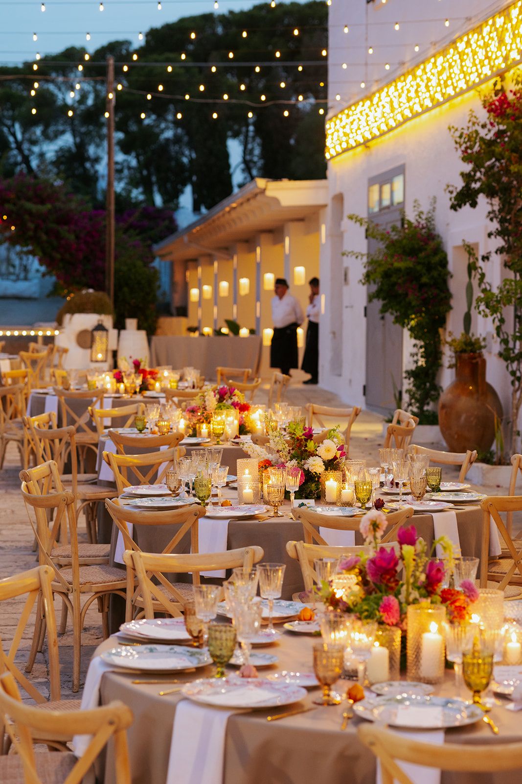Elegant outdoor wedding reception setup with candlelit round tables, cross-back chairs, and romantic floral centerpieces at dusk.