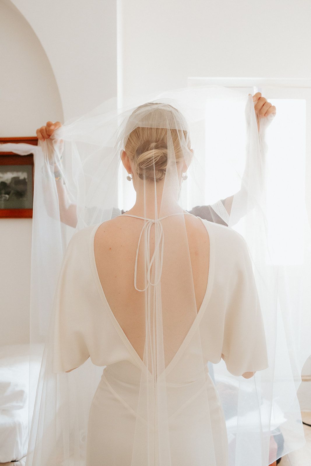 Back view of a bride in a white dress with sheer veil against light wall during wedding preparations.