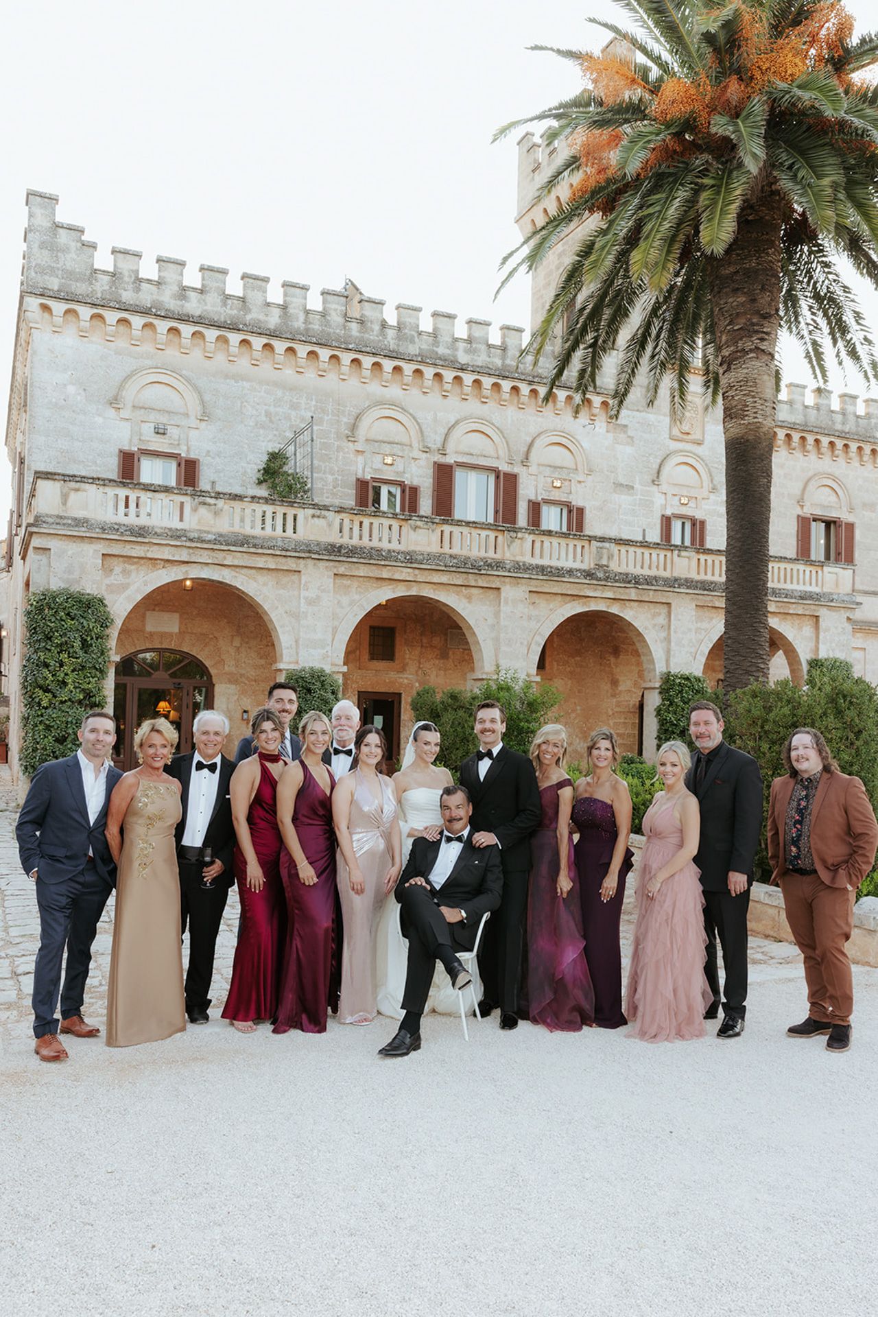 Wedding party in formal attire poses in front of a Mediterranean-style castle with palm trees and stone architecture.