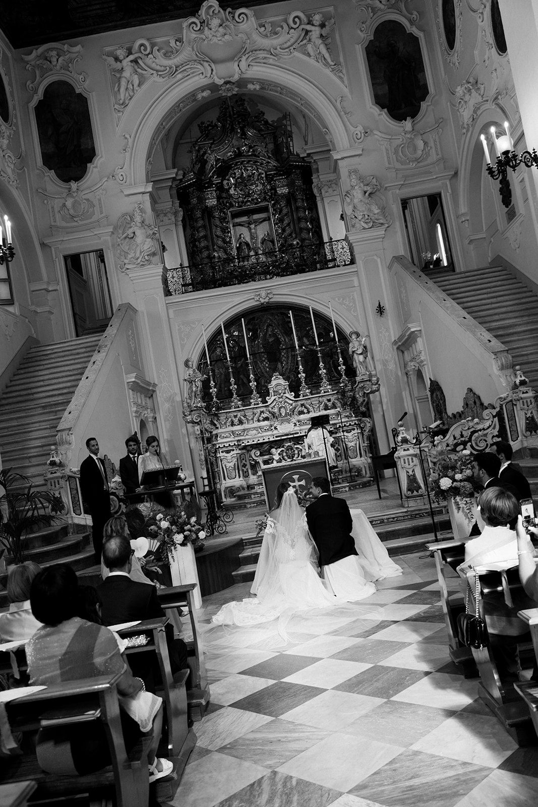 A traditional wedding ceremony taking place inside an ornate Catholic church with a checkered floor and baroque architecture.
