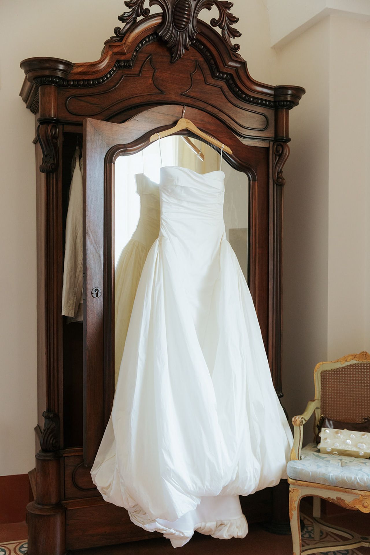 White wedding dress with puffy sleeves displayed against ornate wooden armoire mirrors in vintage setting.