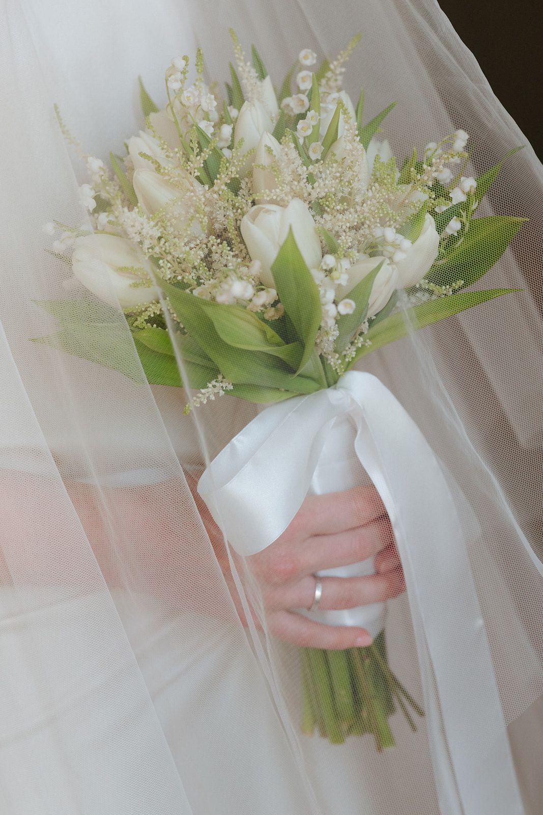 A white floral bouquet with delicate lily of the valley flowers and green leaves held against a soft tulle background.