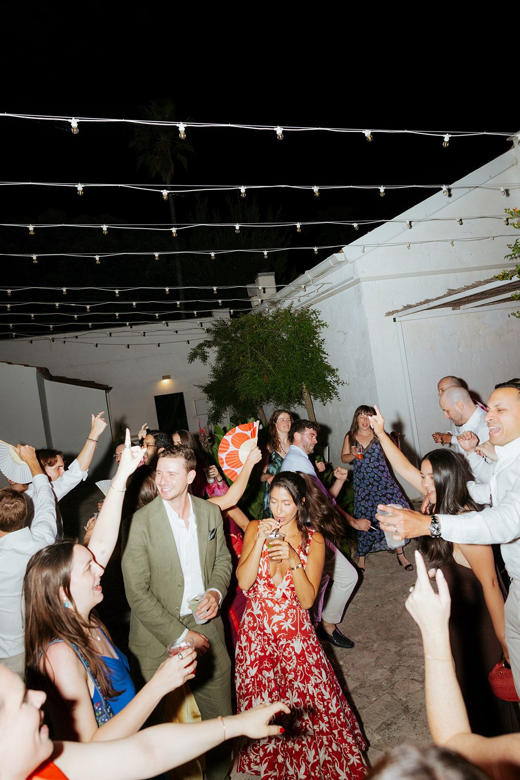 A crowd of wedding guests dancing together with their arms raised under string lights during an evening reception.