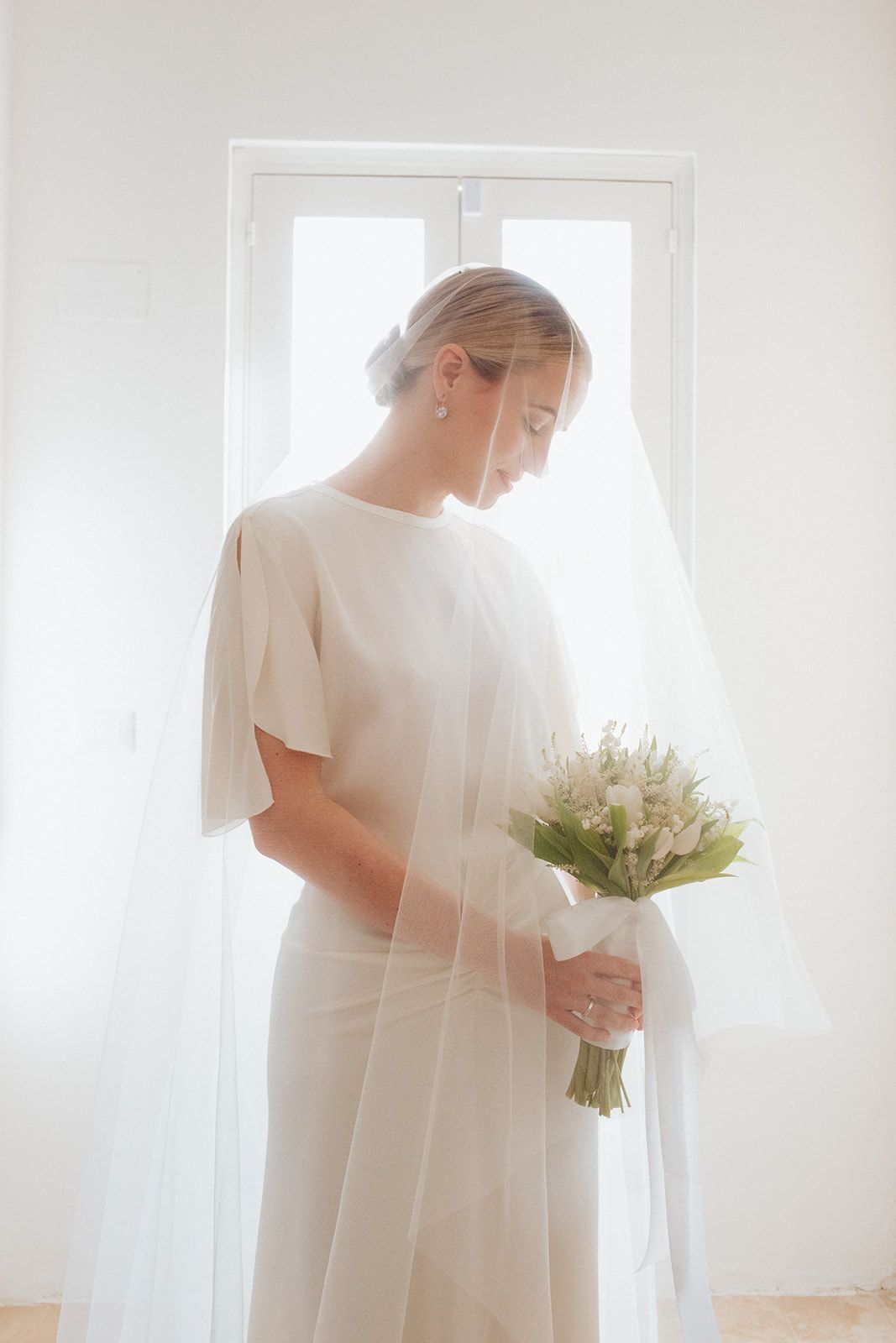 Ethereal bridal portrait with white dress and lily bouquet backlit by window in soft dreamy lighting.